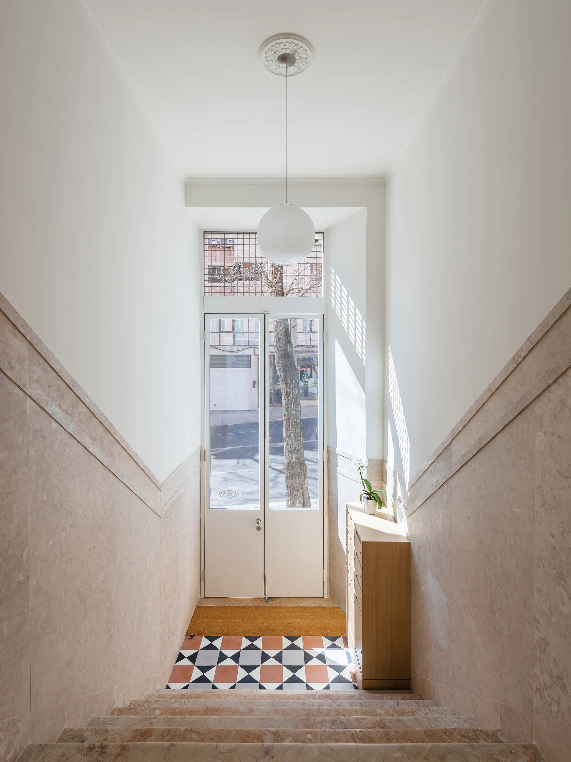 A light-filled entryway stairwell with pinkish stone stairs, matching wall paneling, a geometric tiled landing, and a glass door.