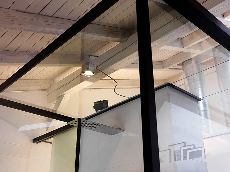 A modern glass shower enclosure with black metal framing stands beneath white-washed exposed wooden ceiling beams and a metallic spotlight.