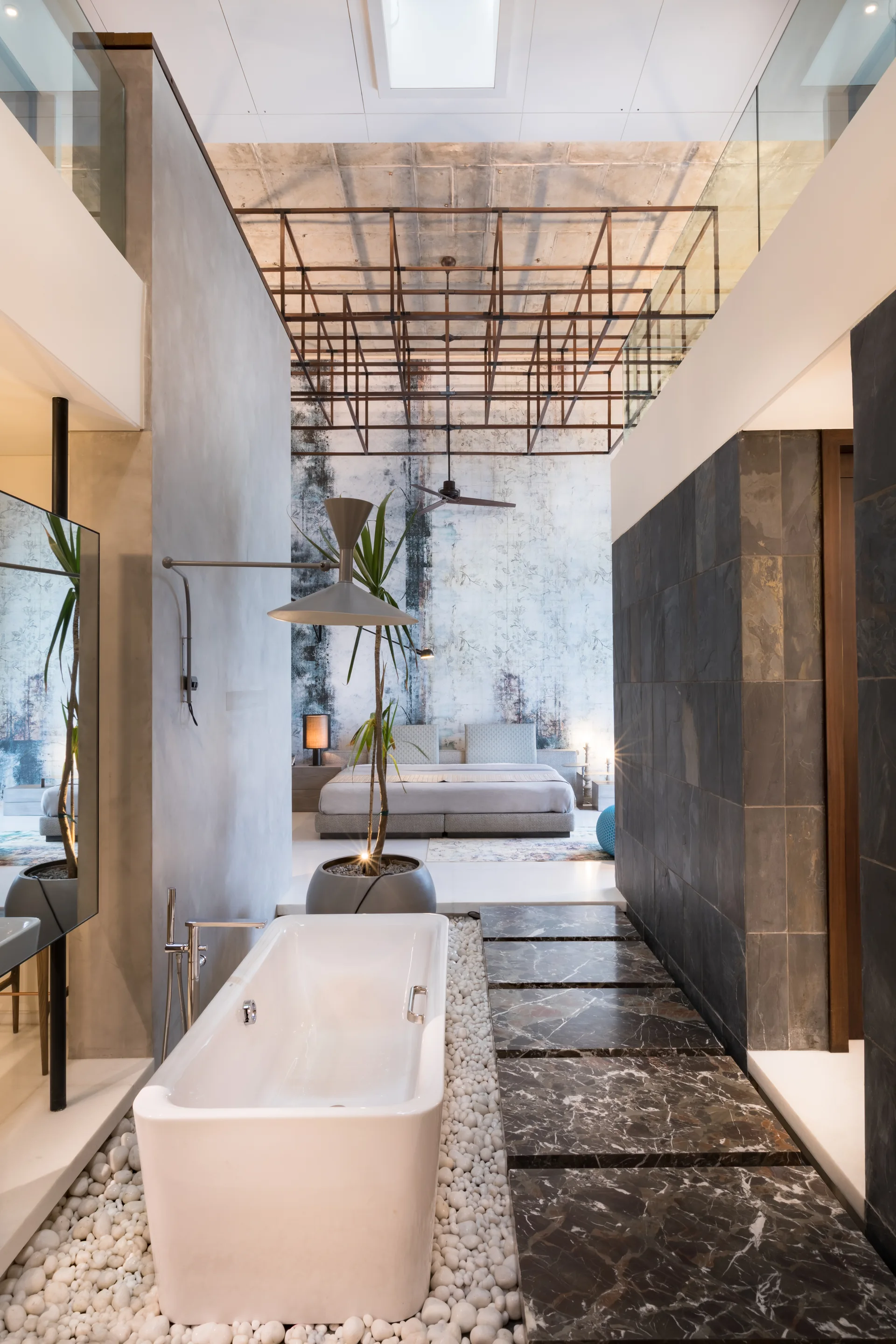 A white freestanding bathtub sits on white pebbles next to dark marble stepping stones in an open-plan en-suite bathroom.