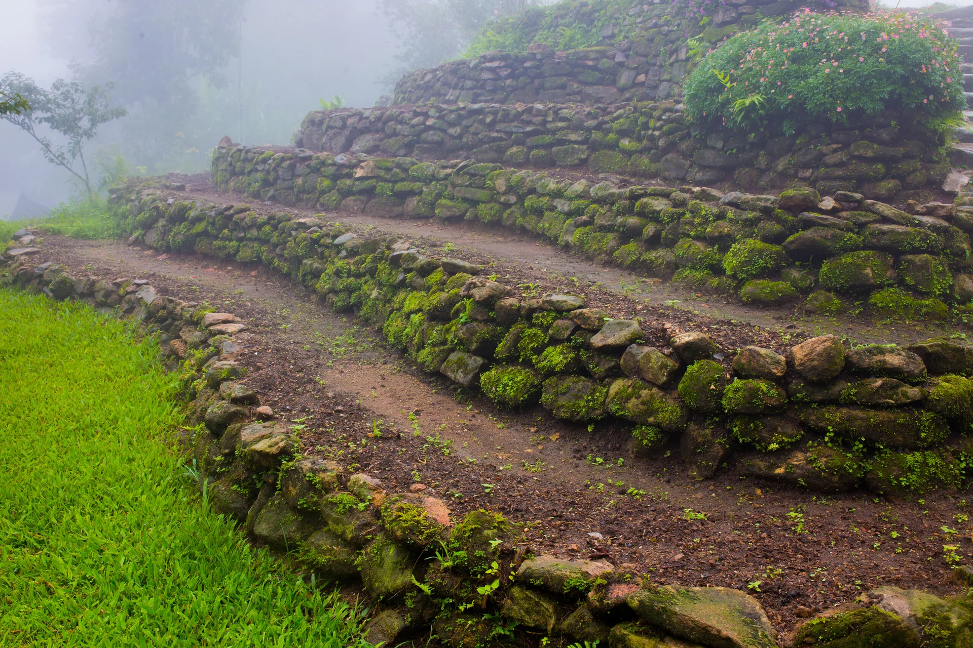 Tiered, moss-covered stone retaining walls rise along a sloped hillside garden with dirt pathways and green grass under misty skies.