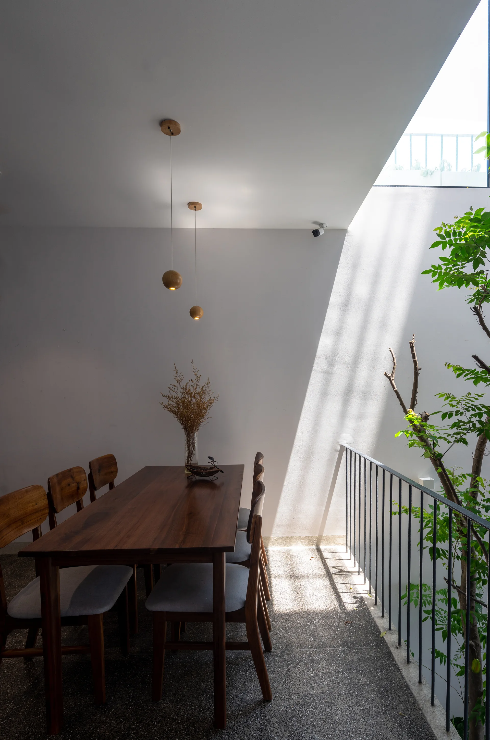 A wooden dining table and chairs sit on a speckled grey floor next to a white wall and a light well with a metal railing and green tree.