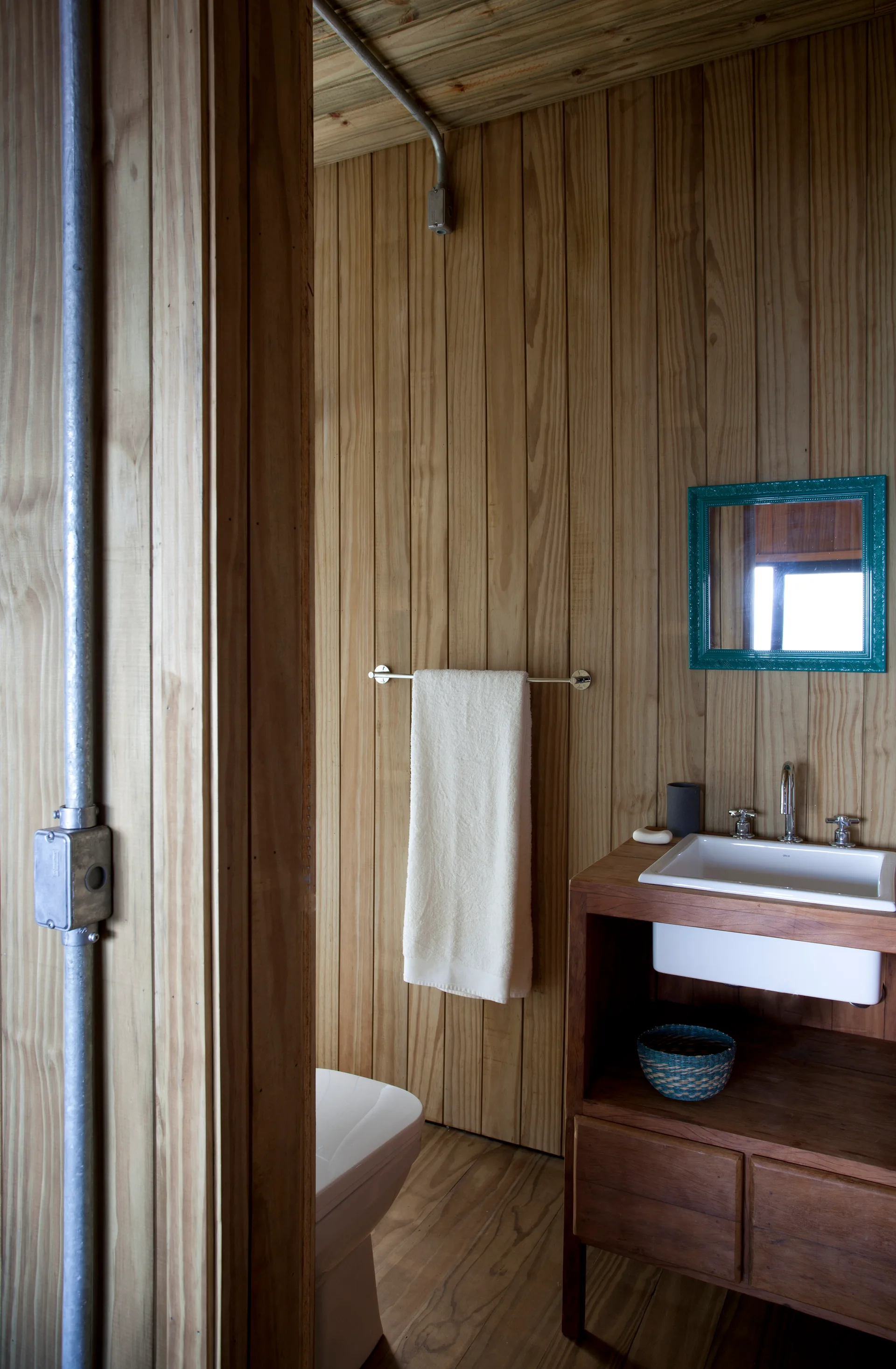 A rustic bathroom with vertical wood plank walls, a wooden vanity with a white sink, a teal mirror frame, and exposed metal conduit.