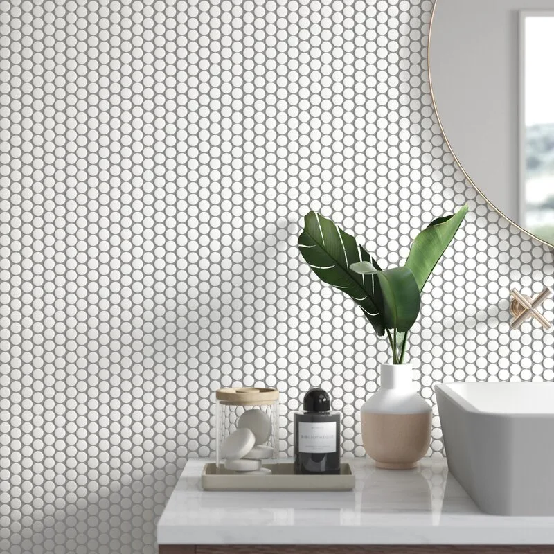 A white penny-tiled bathroom wall behind a marble vanity with a vessel sink, round mirror, and a small plant in a vase.