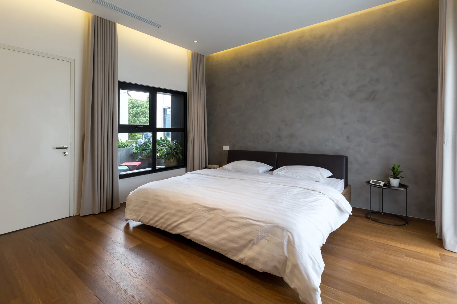 A minimalist bedroom features a bed with white bedding against a textured grey wall, complete with warm wood floors and tall neutral curtains.