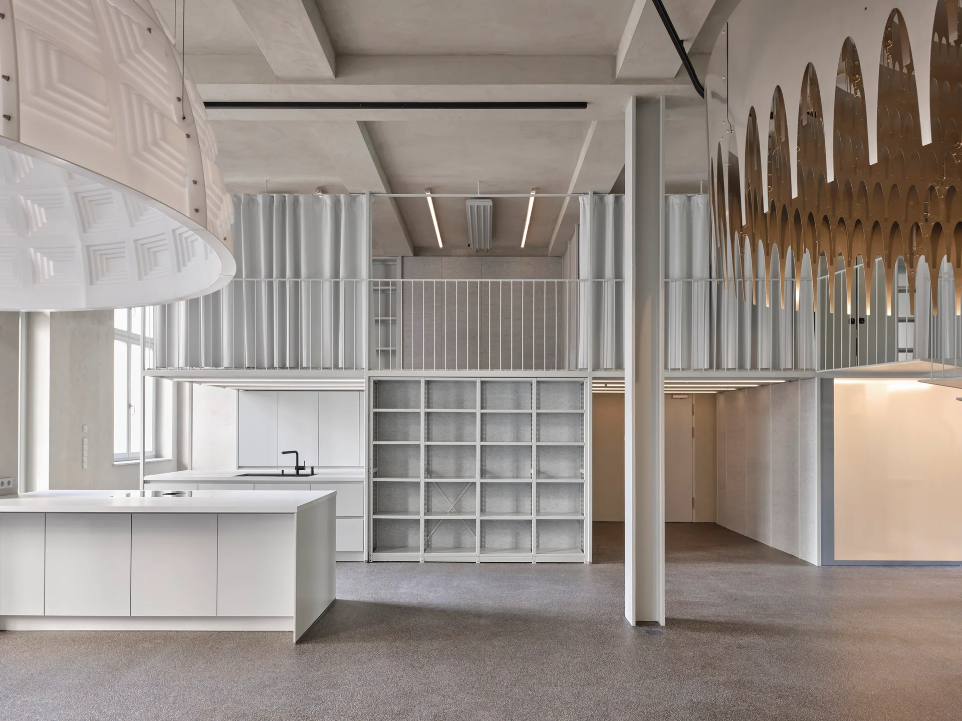 A minimalist white kitchen features a central island beneath a mezzanine level with thin railings, fabric curtains, and an exposed concrete ceiling.