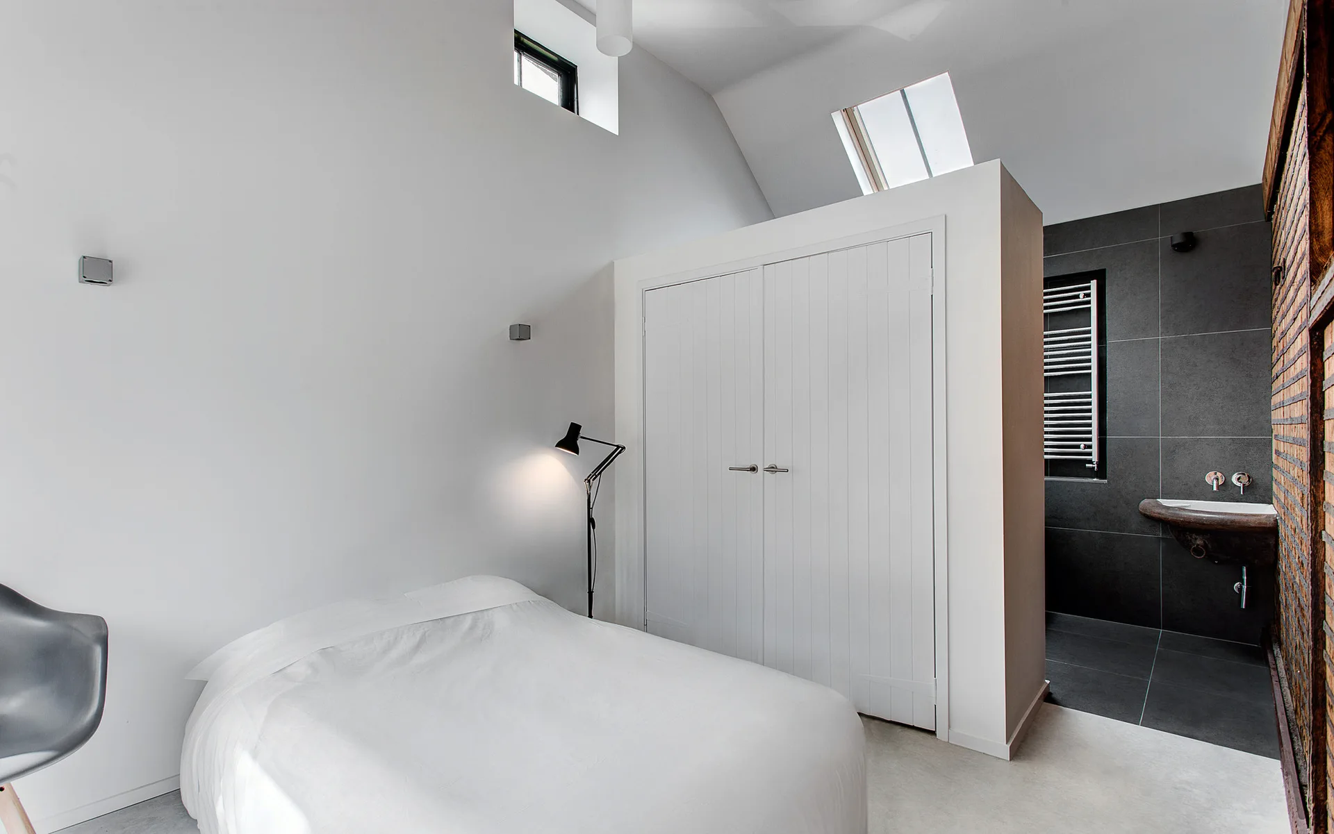 A minimalist bedroom featuring a white bed and a partition wall wardrobe next to a dark-tiled ensuite bathroom.