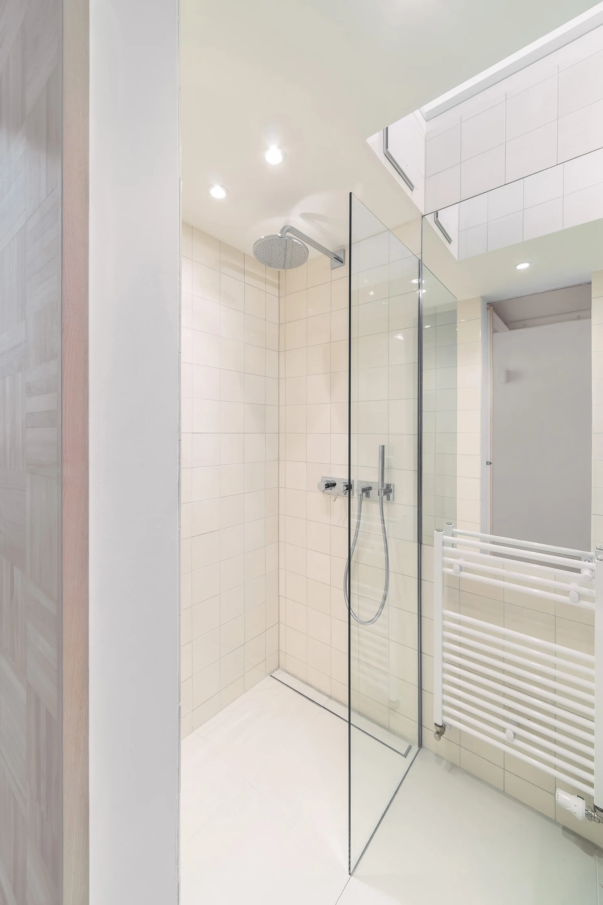 A glass-enclosed walk-in shower featuring cream square wall tiles, a chrome shower head, and an adjacent white ladder-style towel warmer.