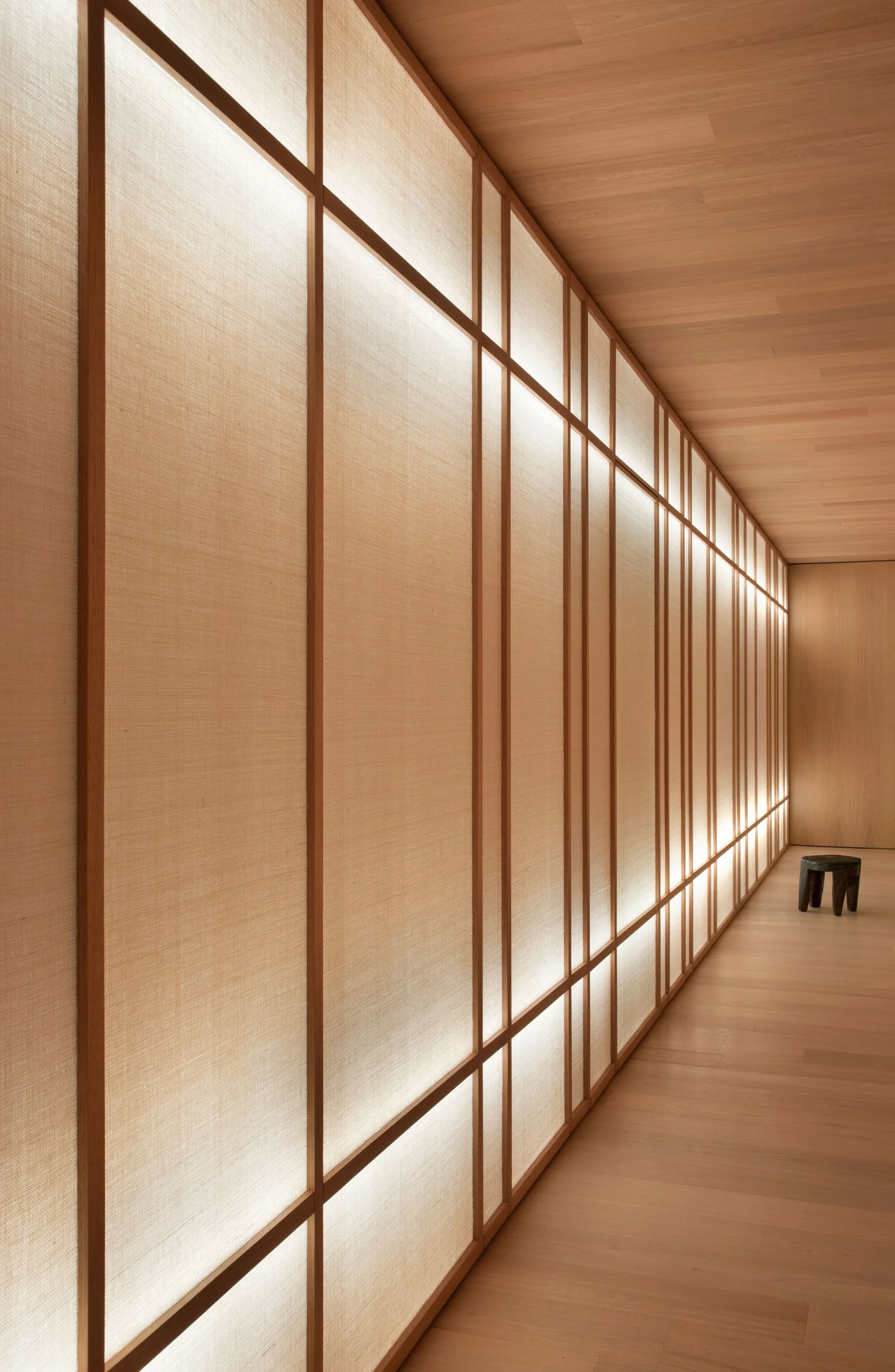 A long corridor features light wood floors and ceilings alongside a glowing, backlit wooden grid partition wall with a small dark stool at the end.