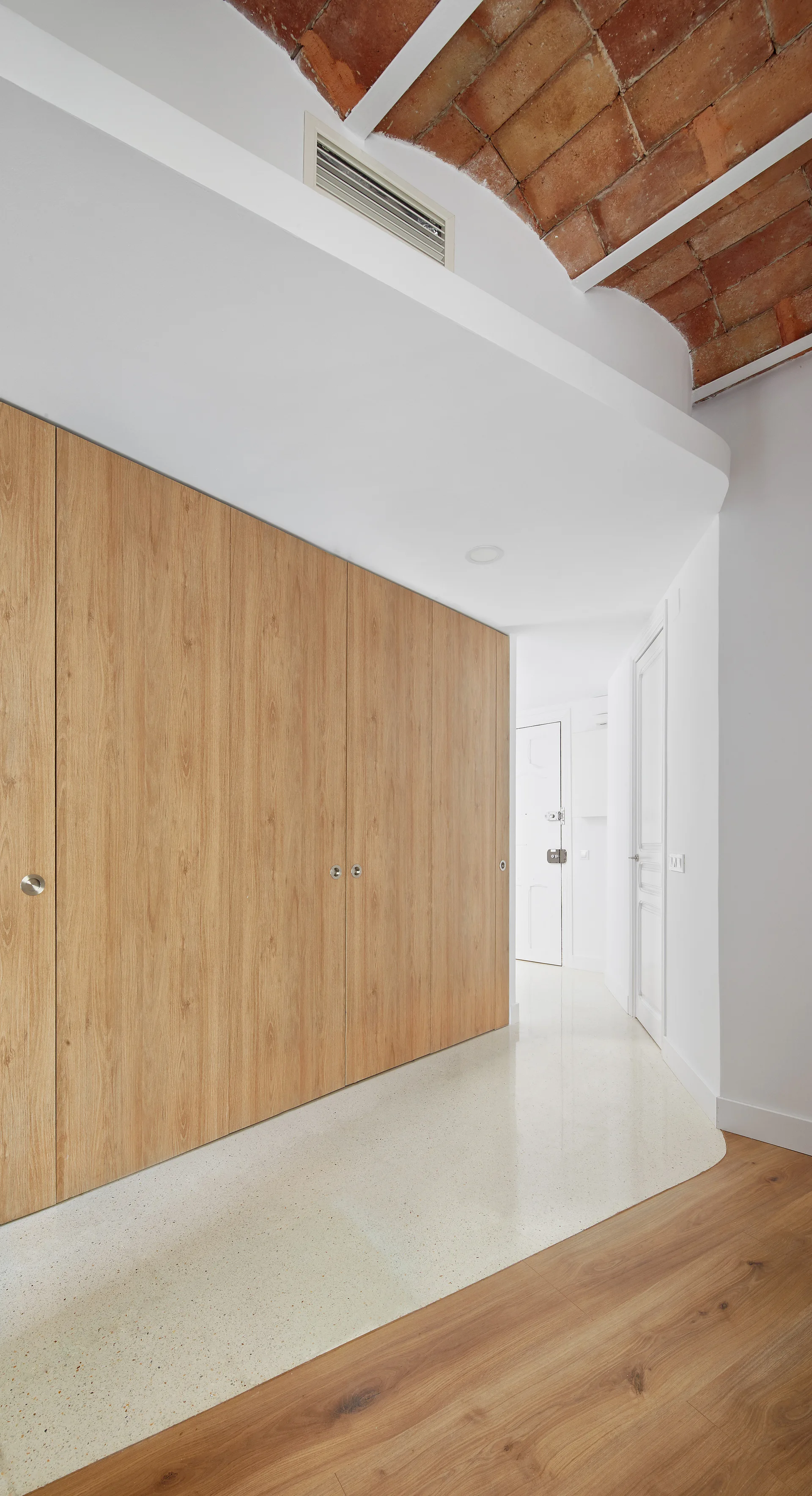 A minimalist hallway featuring integrated light wood cabinets, white walls, and a curved floor transition under an exposed brick vaulted ceiling.