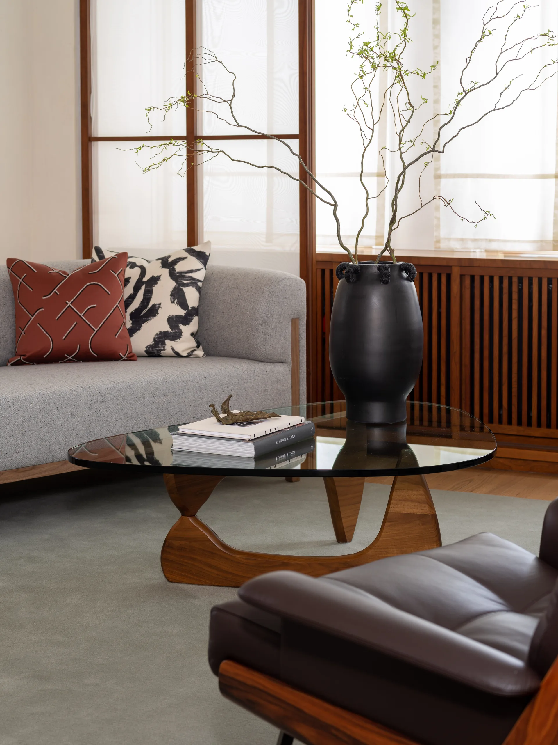 A light grey sofa and dark leather armchair sit near a sculptural glass coffee table holding books and a tall black vase with branches.