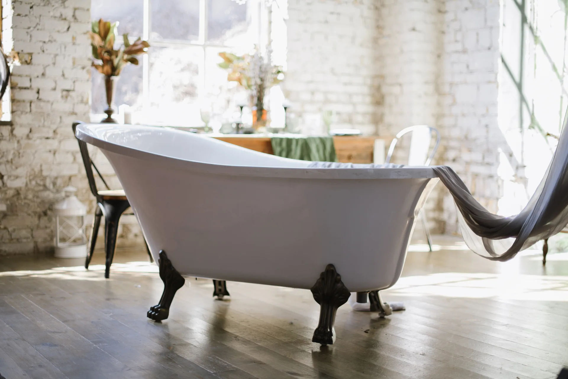 A white freestanding clawfoot bathtub sits on wood floors in a bright loft room with painted brick walls and a large window.