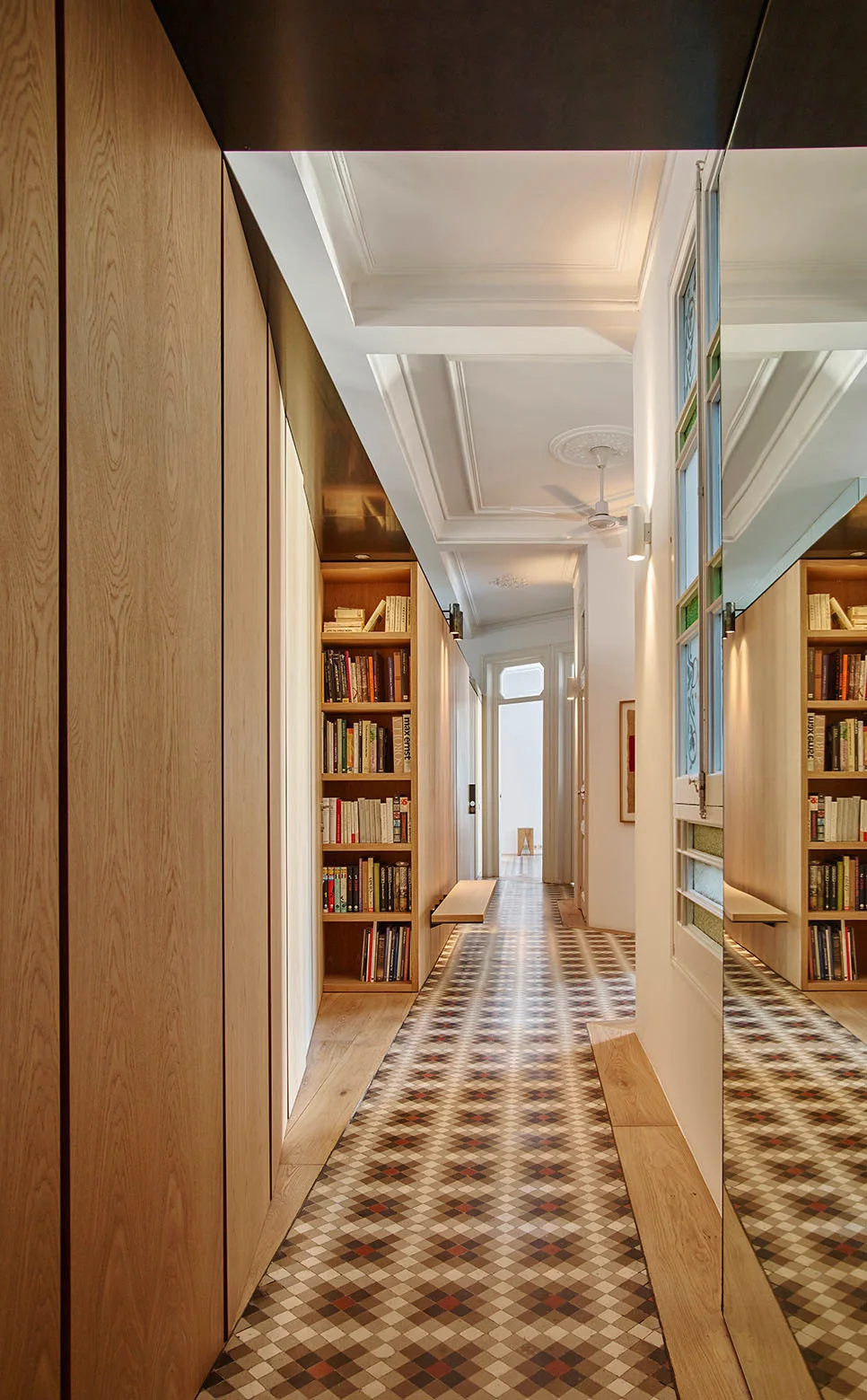 A long hallway features light wood built-in cabinets, a recessed bookshelf, a full-height mirror, and geometric mosaic floor tiles.