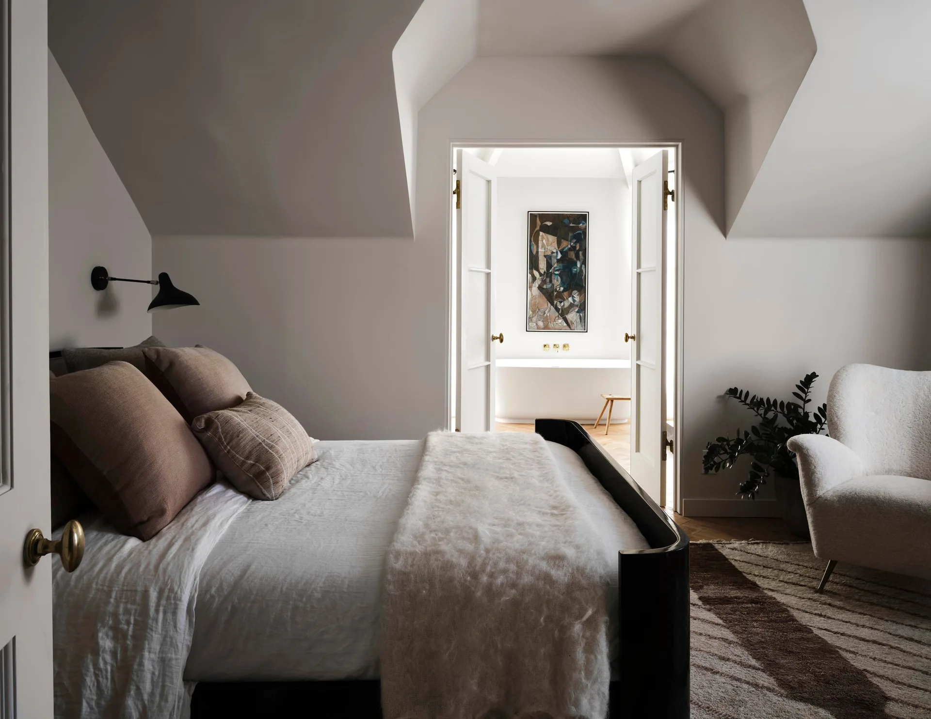 A transitional bedroom with a sloped ceiling, dark bed frame, and open double doors showing a bright bathroom with a freestanding tub.