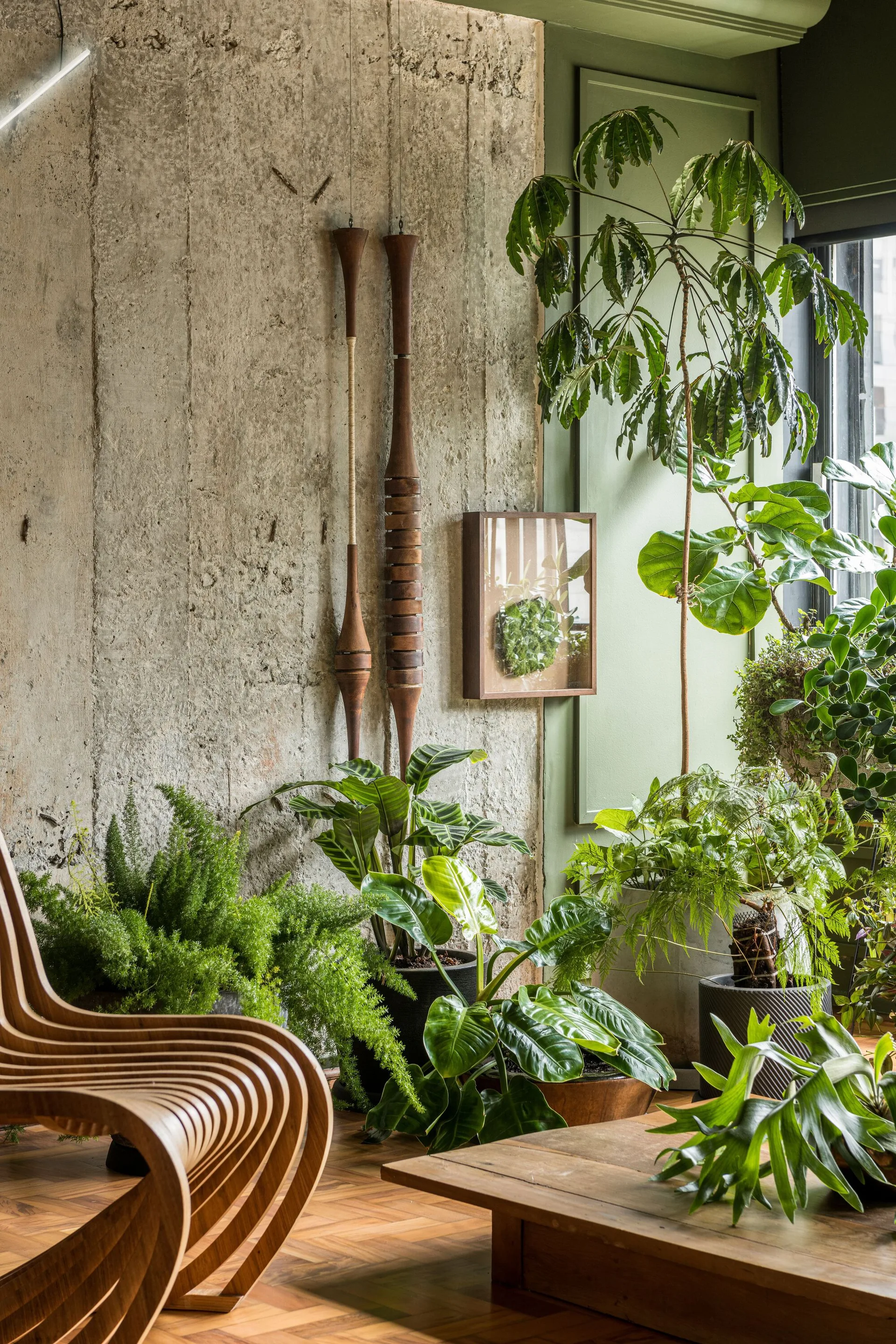 A curved wood slat chair sits in a room with a textured concrete wall, sage green accents, herringbone flooring, and numerous lush houseplants.