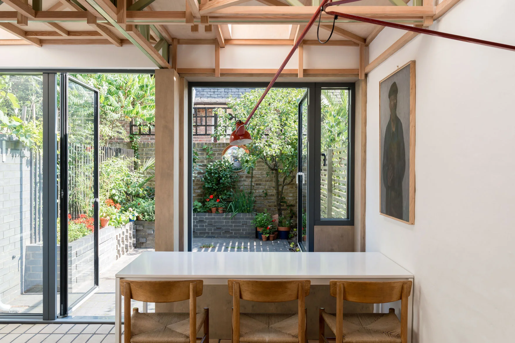 A minimalist dining room with a white table and wooden chairs features an exposed timber ceiling and large glass doors opening to a garden.