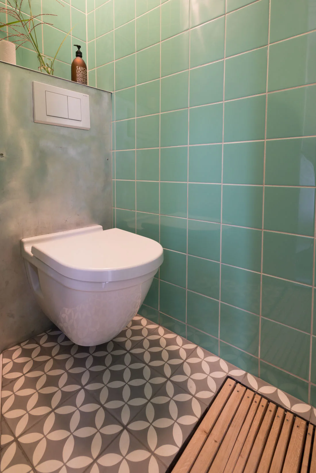 A wall-mounted white toilet stands against glossy mint green square wall tiles above a grey and white geometric patterned tile floor.