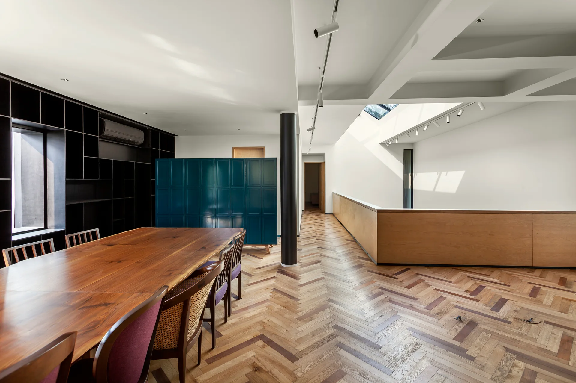 A dining area with a long wood table and chairs next to a teal cabinet and a hallway with herringbone wood floors.
