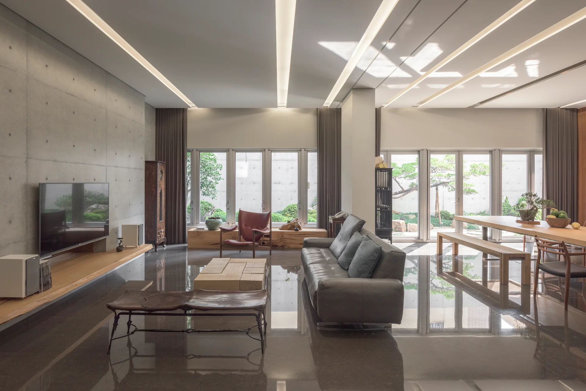 A spacious living room with polished dark reflective floors, concrete panel walls, a grey leather sofa, and linear recessed ceiling lights.