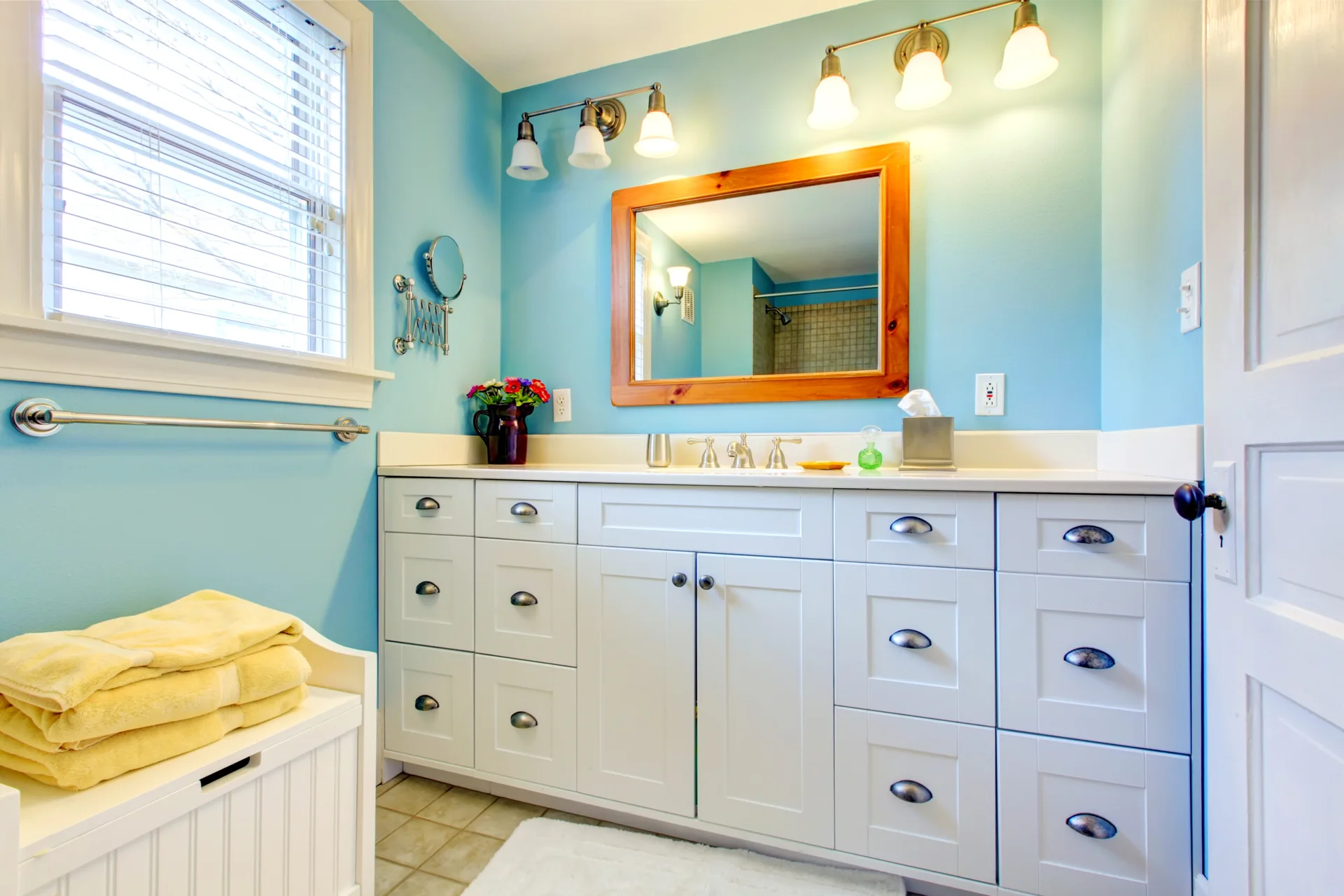 A light blue bathroom features a long white vanity with shaker-style cabinets, a natural wood-framed mirror, and warm multi-bulb light fixtures.