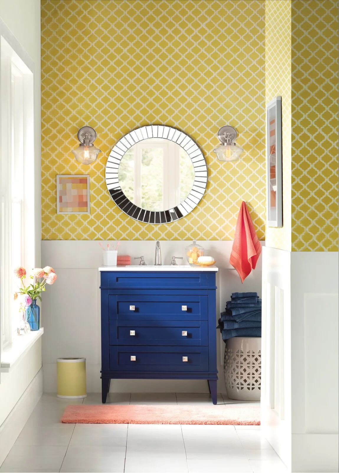A bold blue vanity stands against yellow patterned wallpaper and white wall paneling in a bright bathroom with a circular mirror.