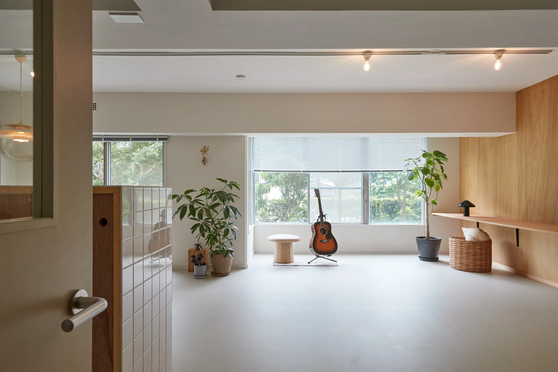 A minimalist living room features a light wood paneled wall, a long floating shelf, potted plants, and an acoustic guitar centered in front of a window.