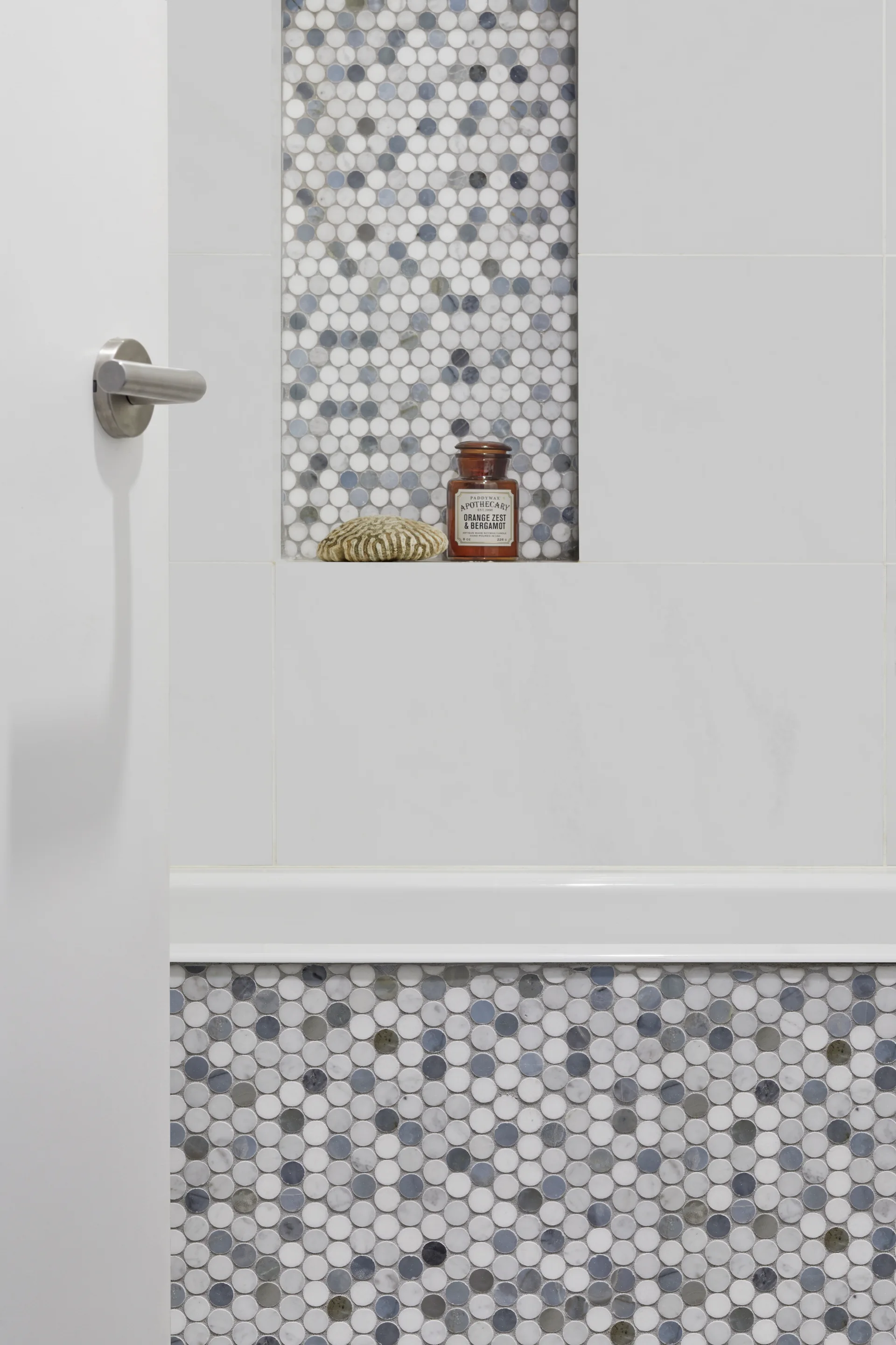 A modern bathroom wall features large white tiles, a recessed niche lined with grey and white penny round mosaic tile, and matching lower wall tiling.