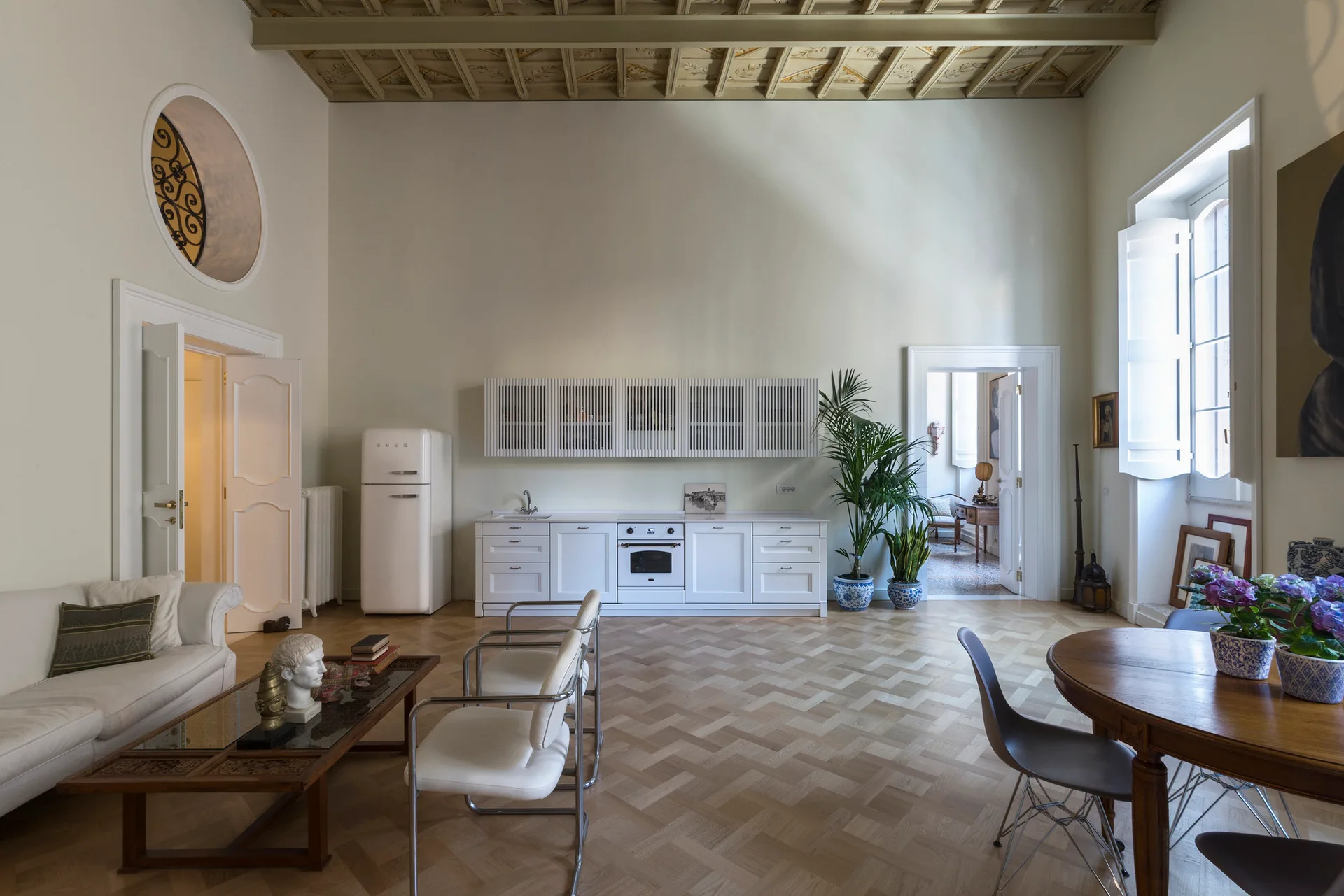 An open living space features a minimalist white kitchen line, a retro refrigerator, a coffered wood ceiling, and herringbone parquet floors.
