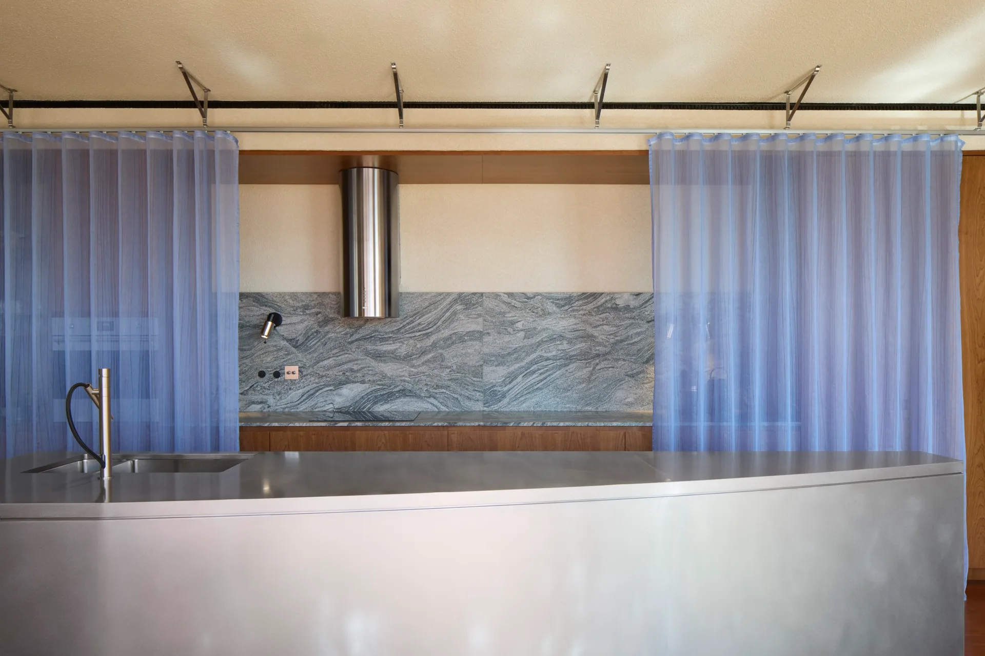 A minimalist kitchen features a stainless steel island and veined grey marble backsplash framed by sheer blue curtains hanging from ceiling tracks.