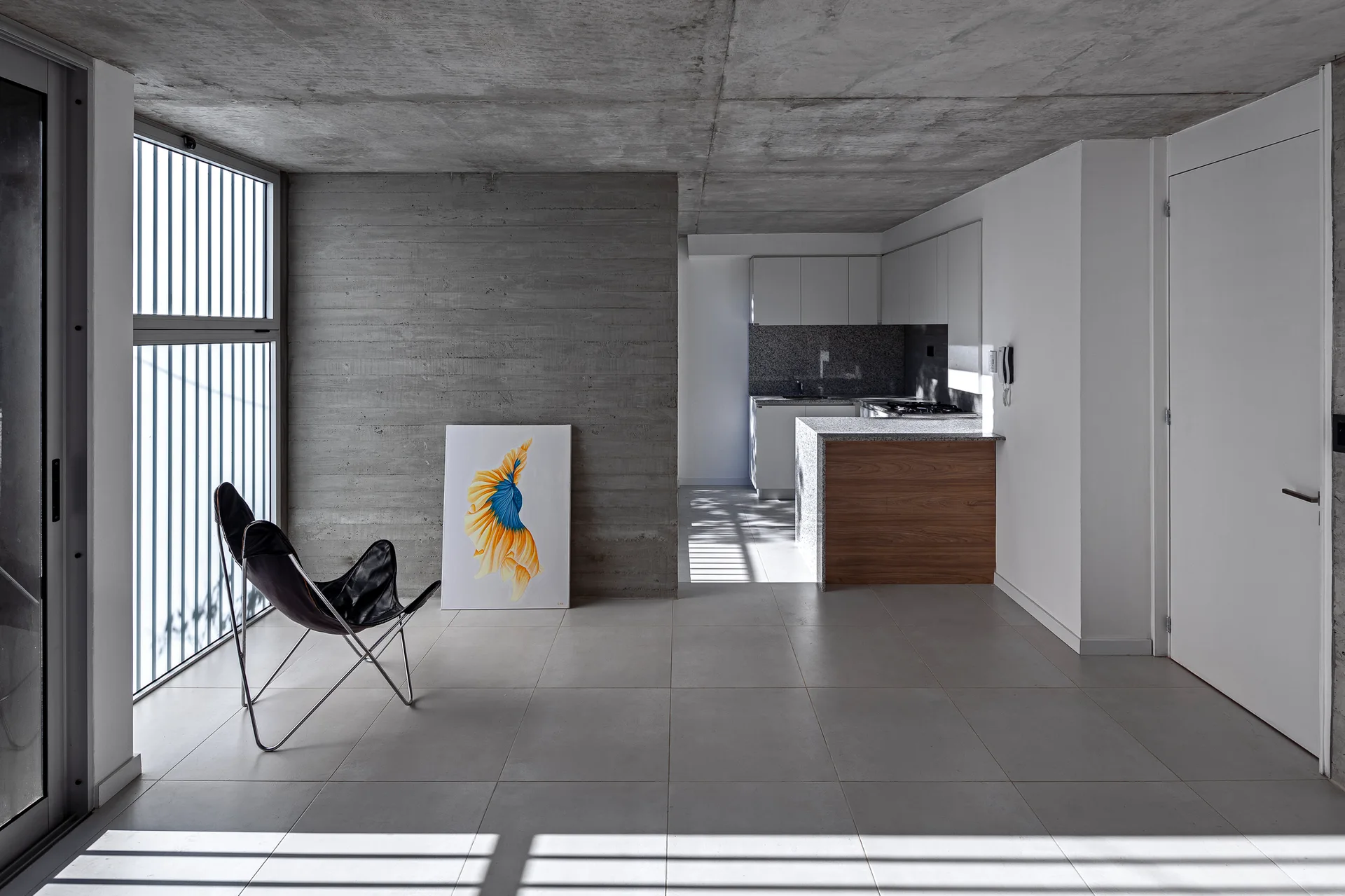 A minimalist modern interior features a black leather butterfly chair, a board-formed concrete wall, and a wood-accented kitchen island in the background.