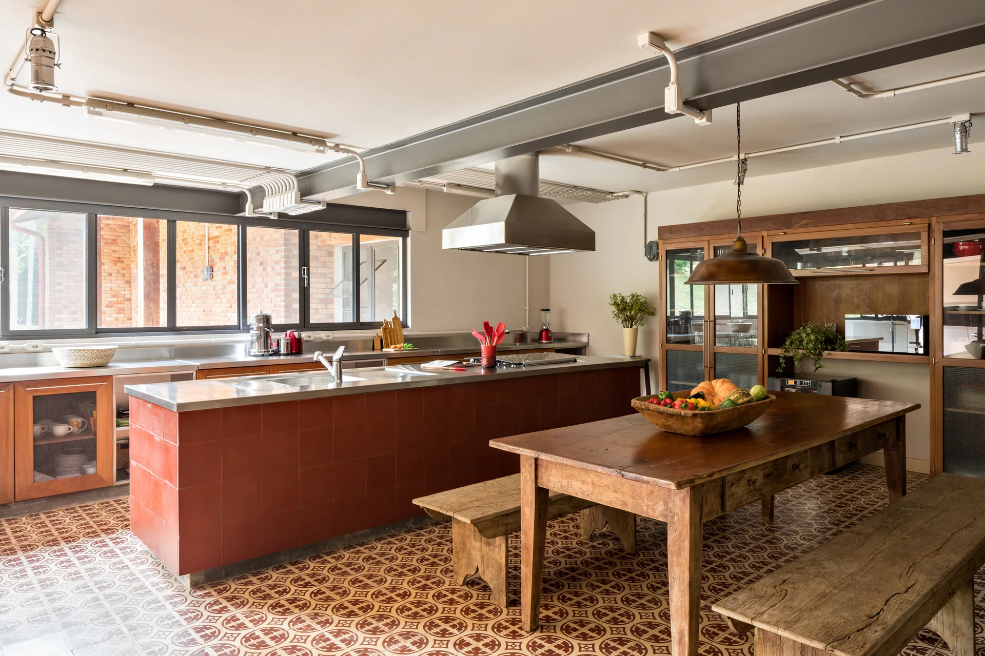 An industrial kitchen features a red tiled island with stainless steel counters, a rustic wooden dining table, and patterned floor tiles.