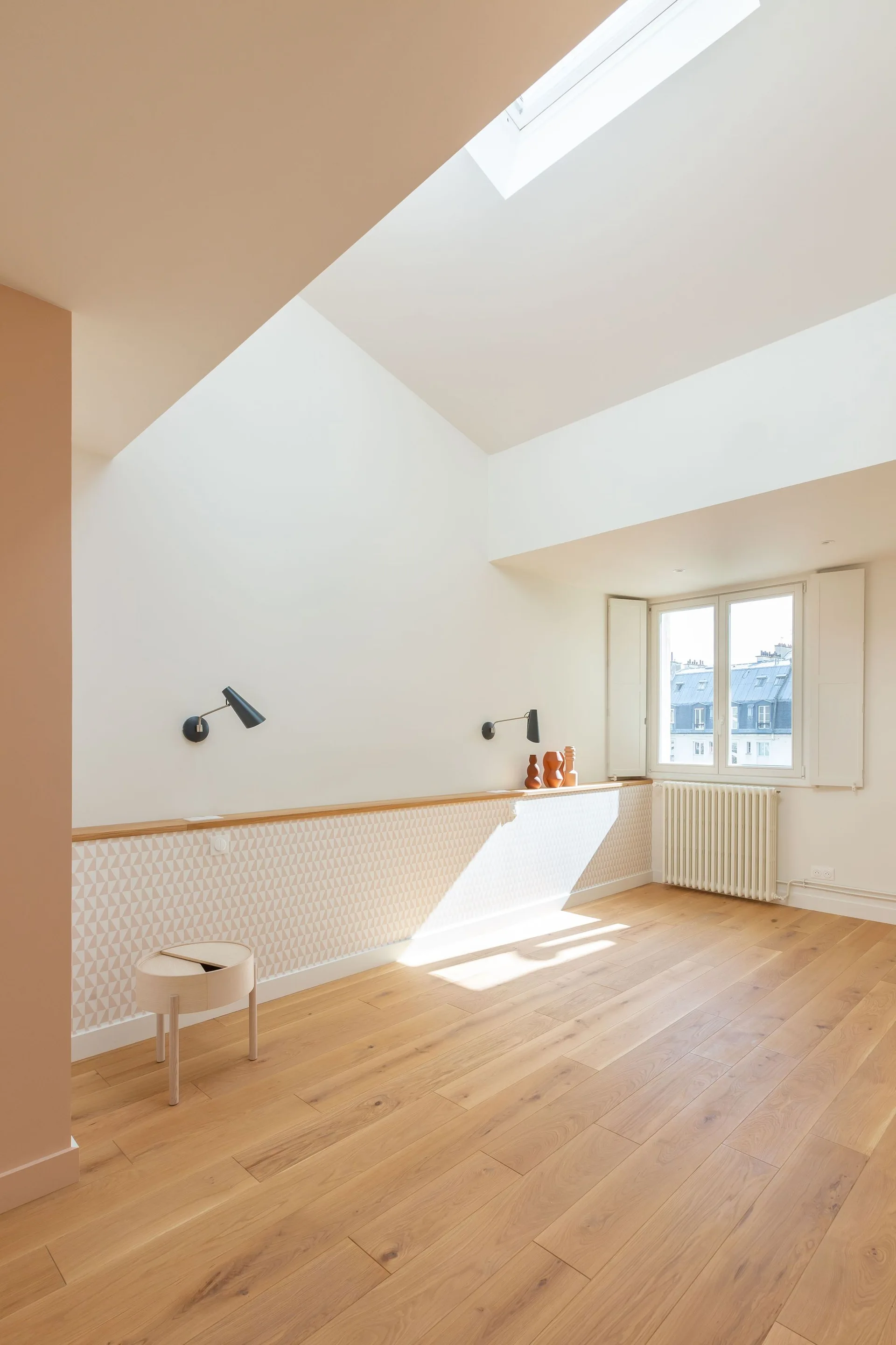 A bright attic bedroom featuring light wood flooring, a sloped ceiling with a skylight, and a long wood-topped ledge with geometric wallpaper.