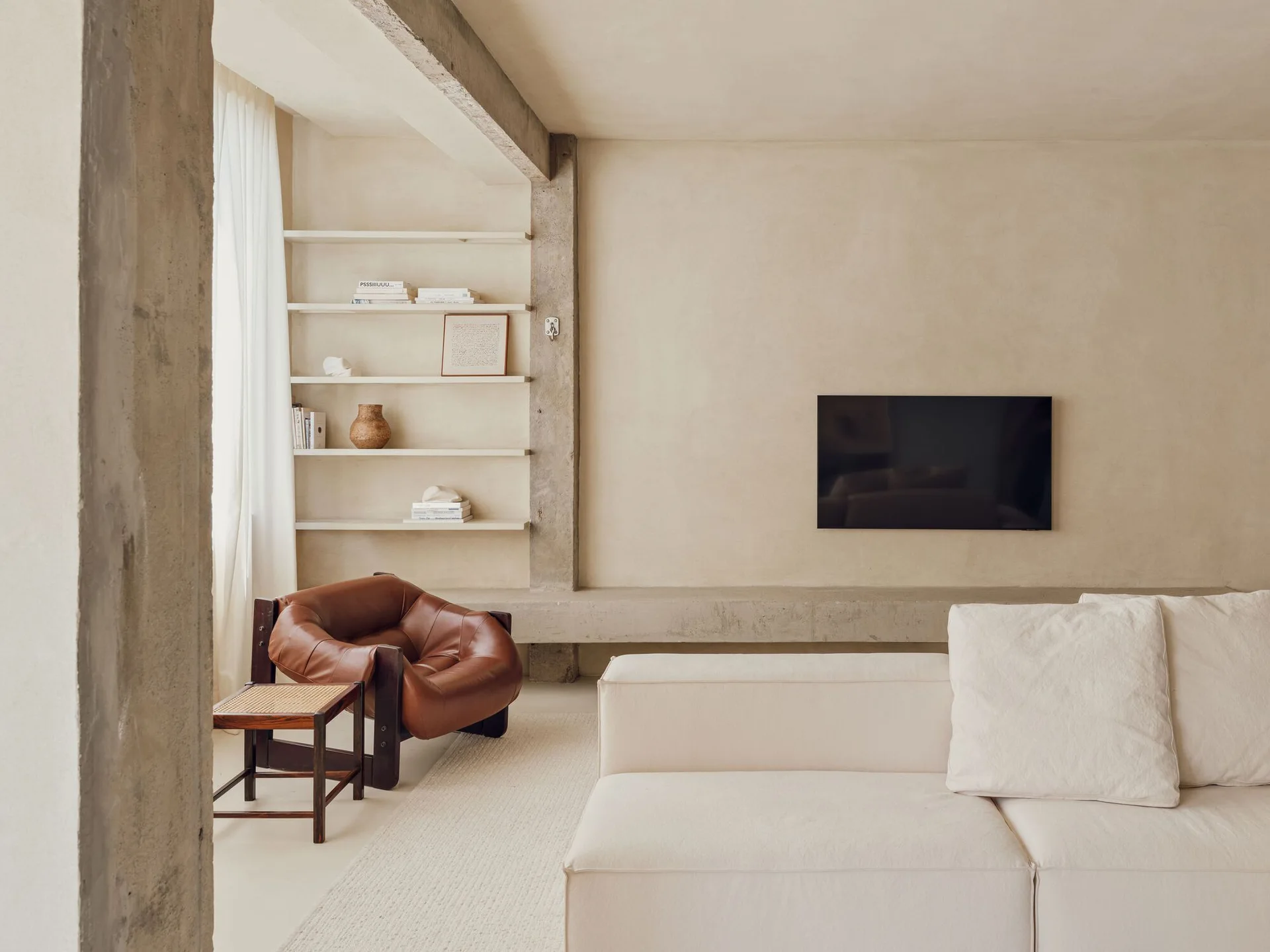 A minimalist living room features a cream plaster wall, an exposed concrete beam, built-in shelving, a white sofa, and a brown leather armchair.