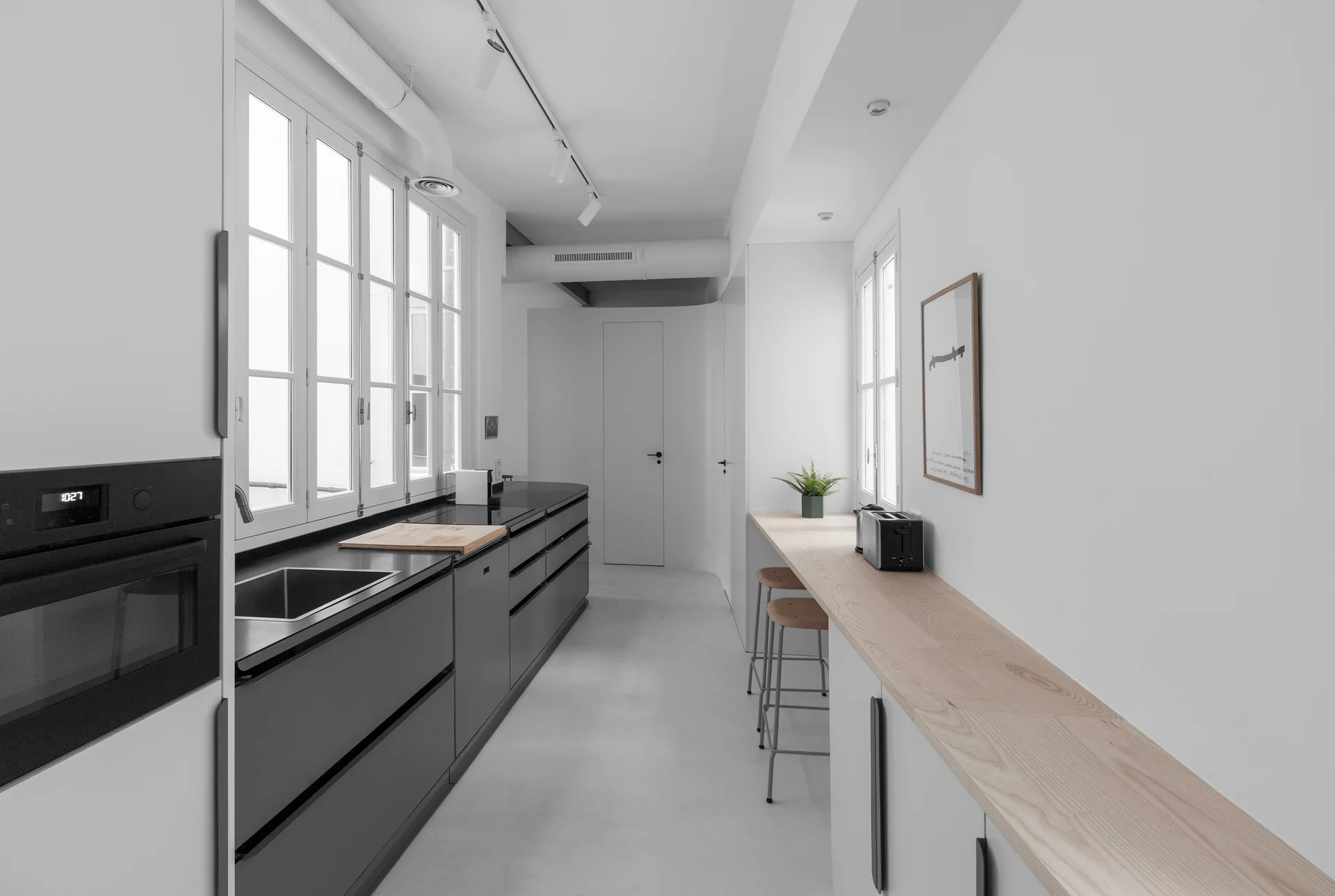 A minimalist galley kitchen featuring dark grey cabinets below large white windows and a long wooden breakfast bar with stools.