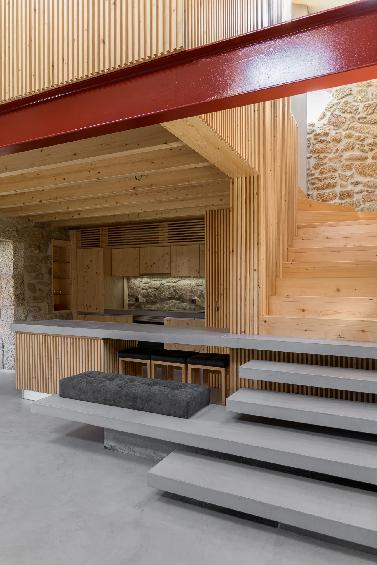 A modern interior featuring a light wood staircase, vertical wood slats, concrete steps, and a prominent red structural steel beam.