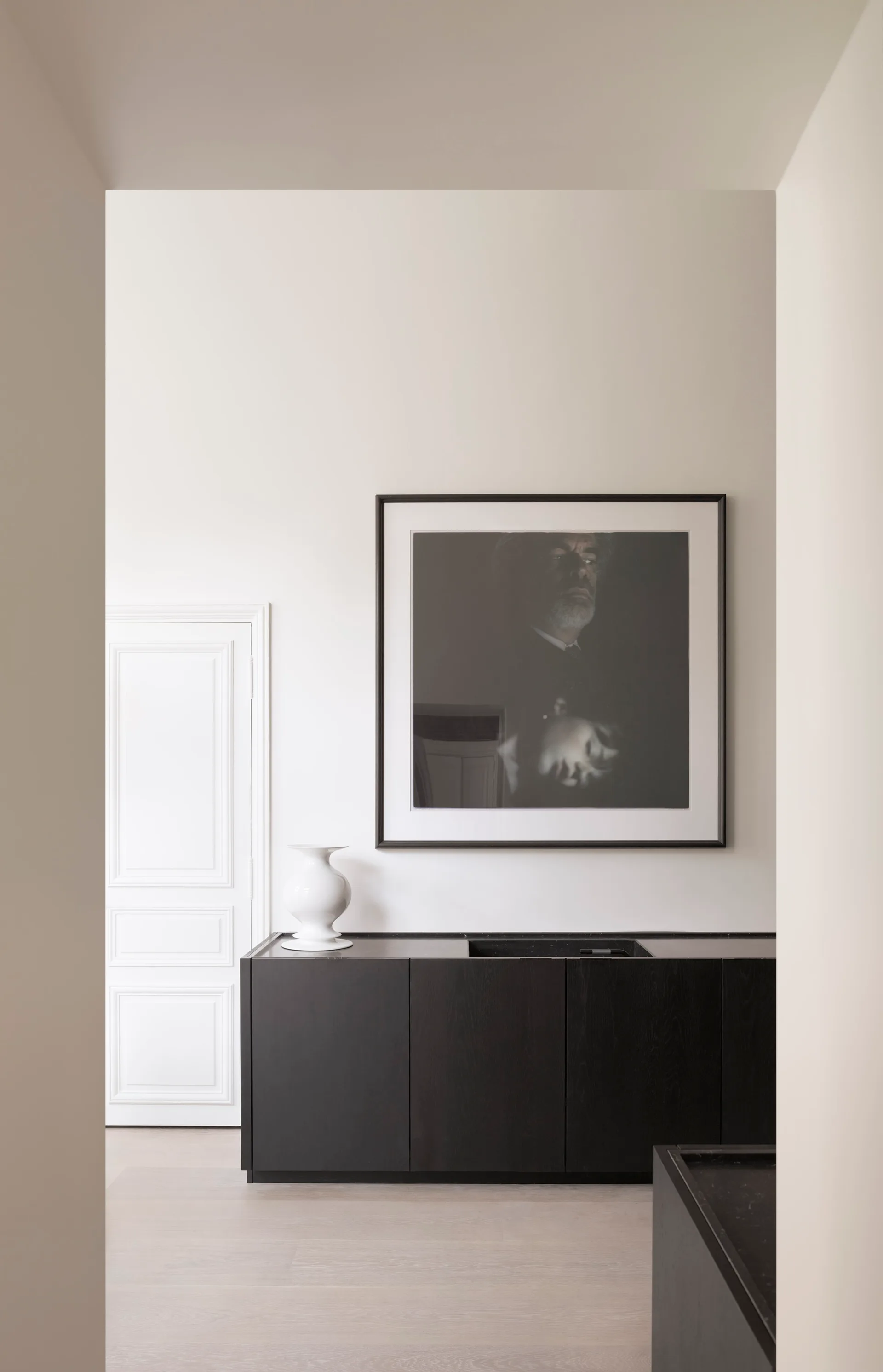 A minimalist hallway features a dark wooden console cabinet, a large dark framed photograph on a white wall, and a classic white paneled door.