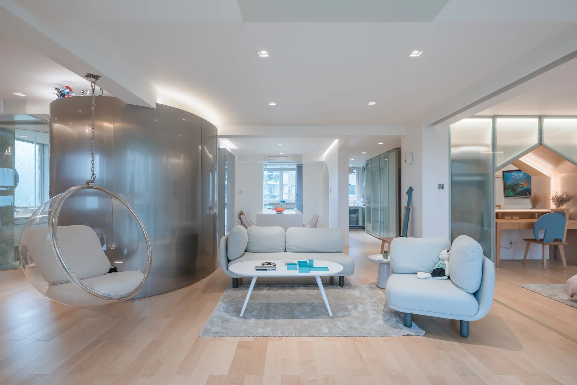 A modern open-concept living room featuring a curved silver partition wall, a hanging acrylic bubble chair, light gray sofas, and blonde wood floors.