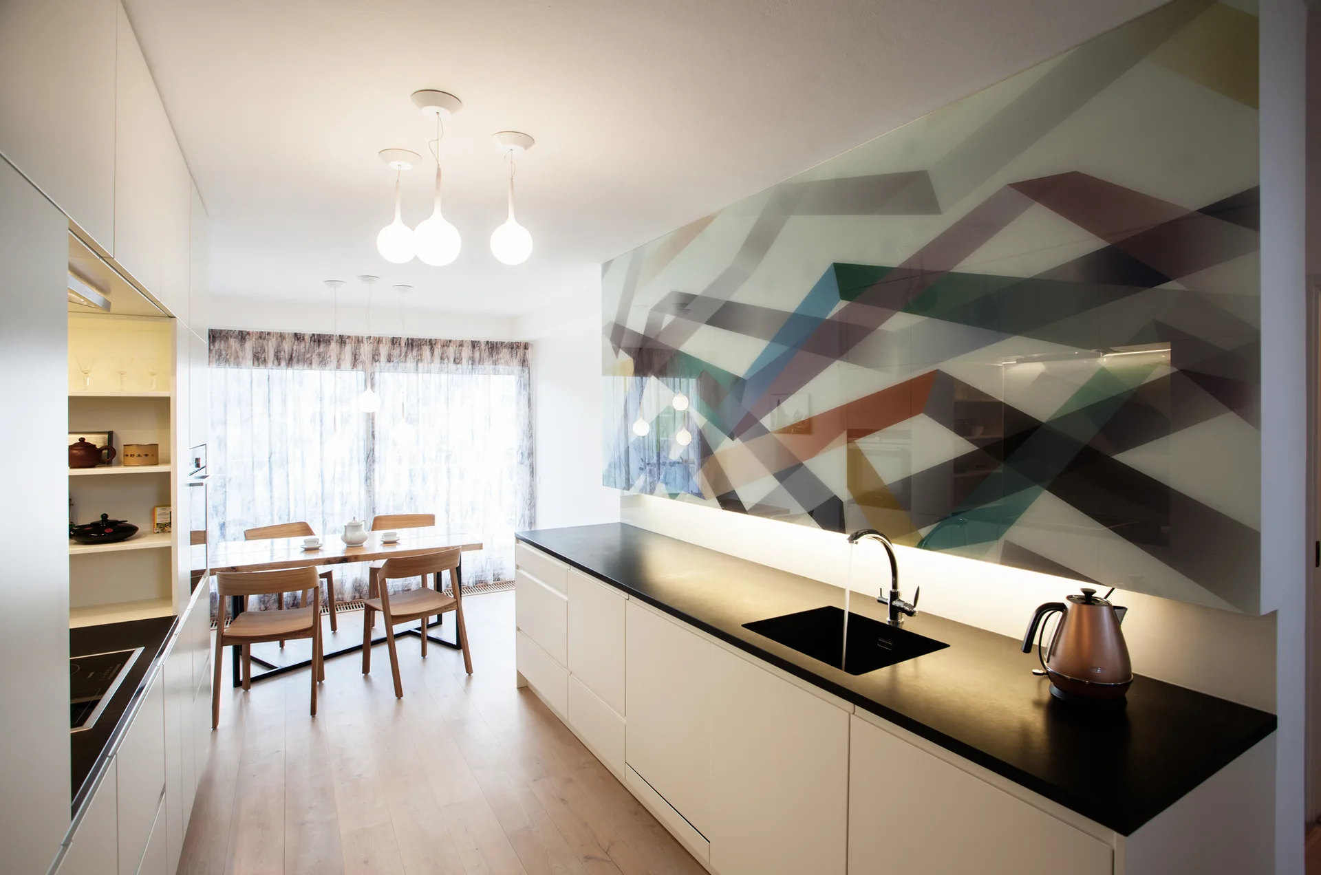 A modern kitchen features white flat-panel cabinets, a black countertop, and a large colorful geometric glass backsplash above the sink.