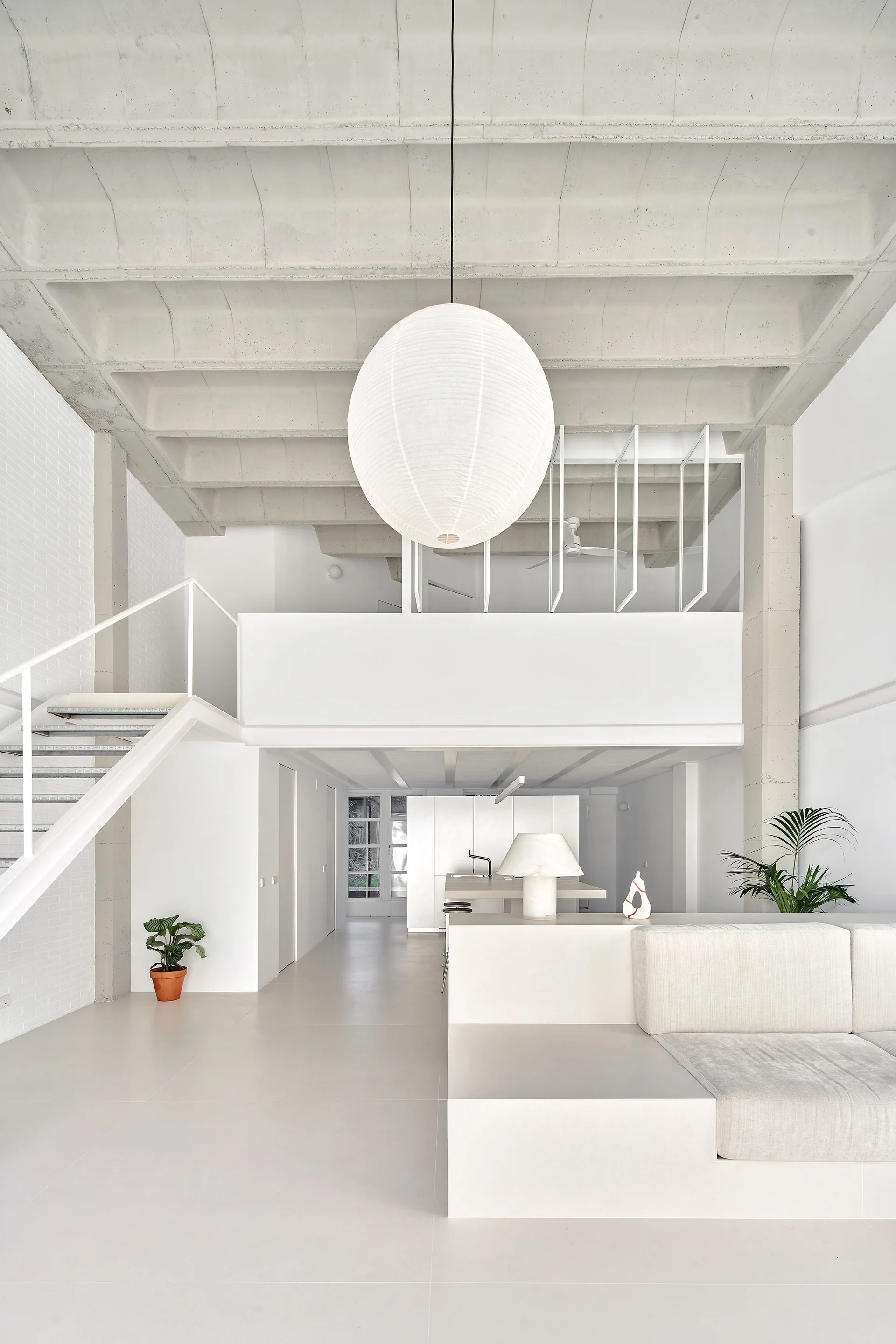 A minimalist white loft living room features a built-in sofa, a metal staircase, and an exposed ribbed concrete ceiling with a large paper lantern.