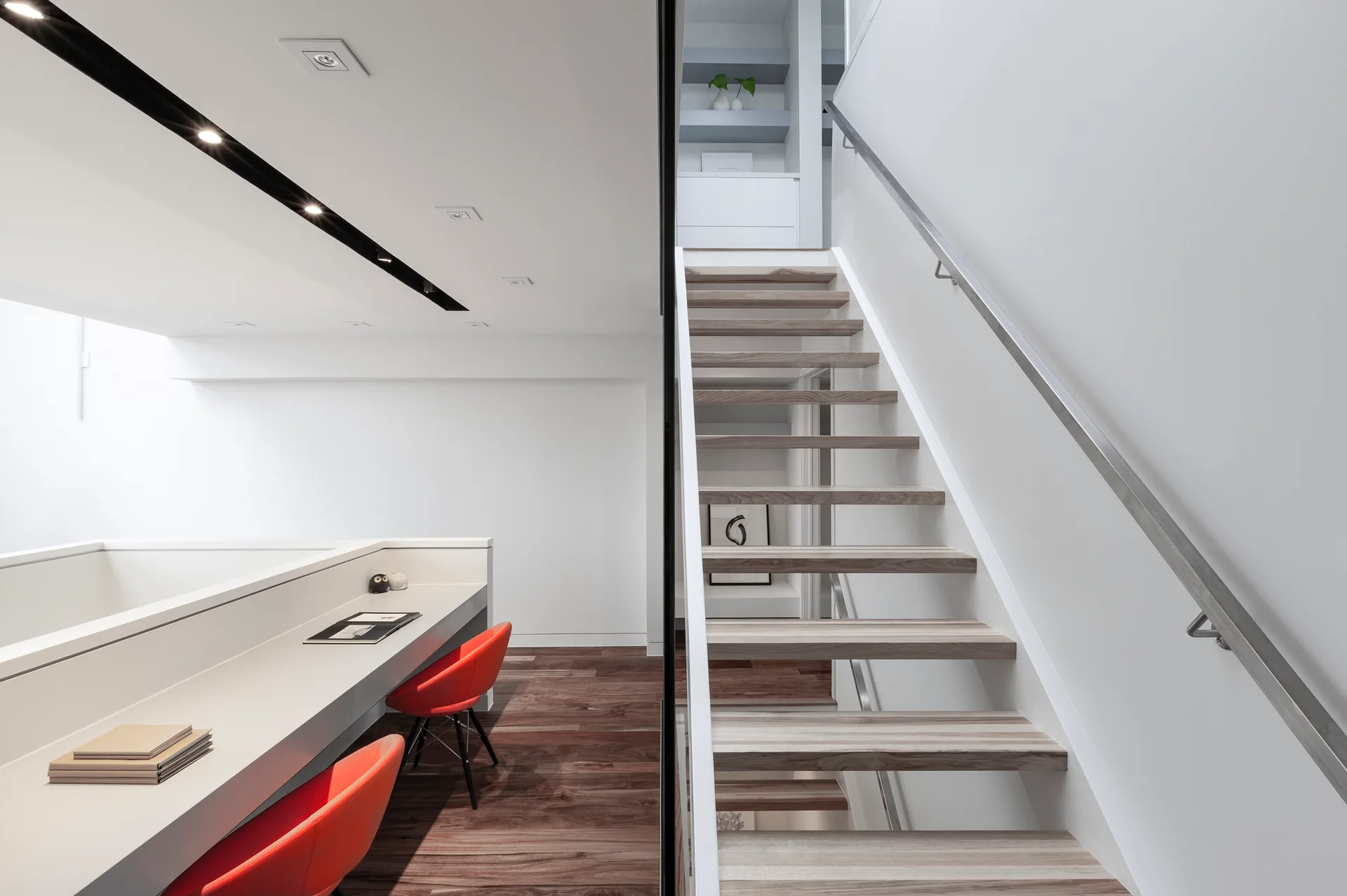 A minimalist modern home office featuring a floating wooden staircase next to a long white built-in desk with red chairs.