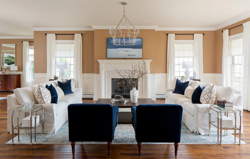 A symmetrical living room featuring two white sofas, two dark blue armchairs, a central white fireplace, and tan walls with white wainscoting.