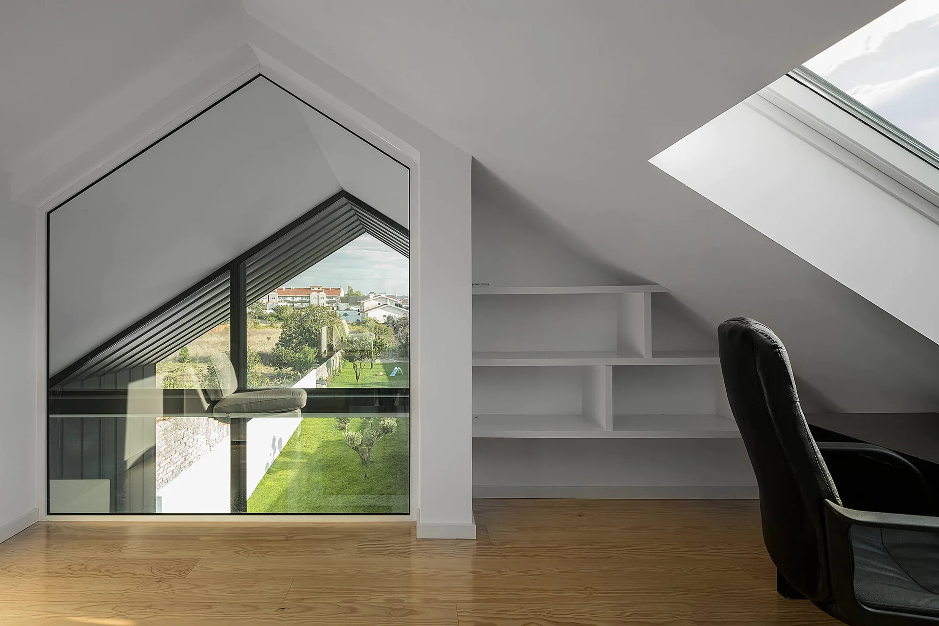 A minimalist attic workspace featuring a sloped ceiling, light wood floors, built-in white shelving, and a large window looking out onto a grassy landscape.