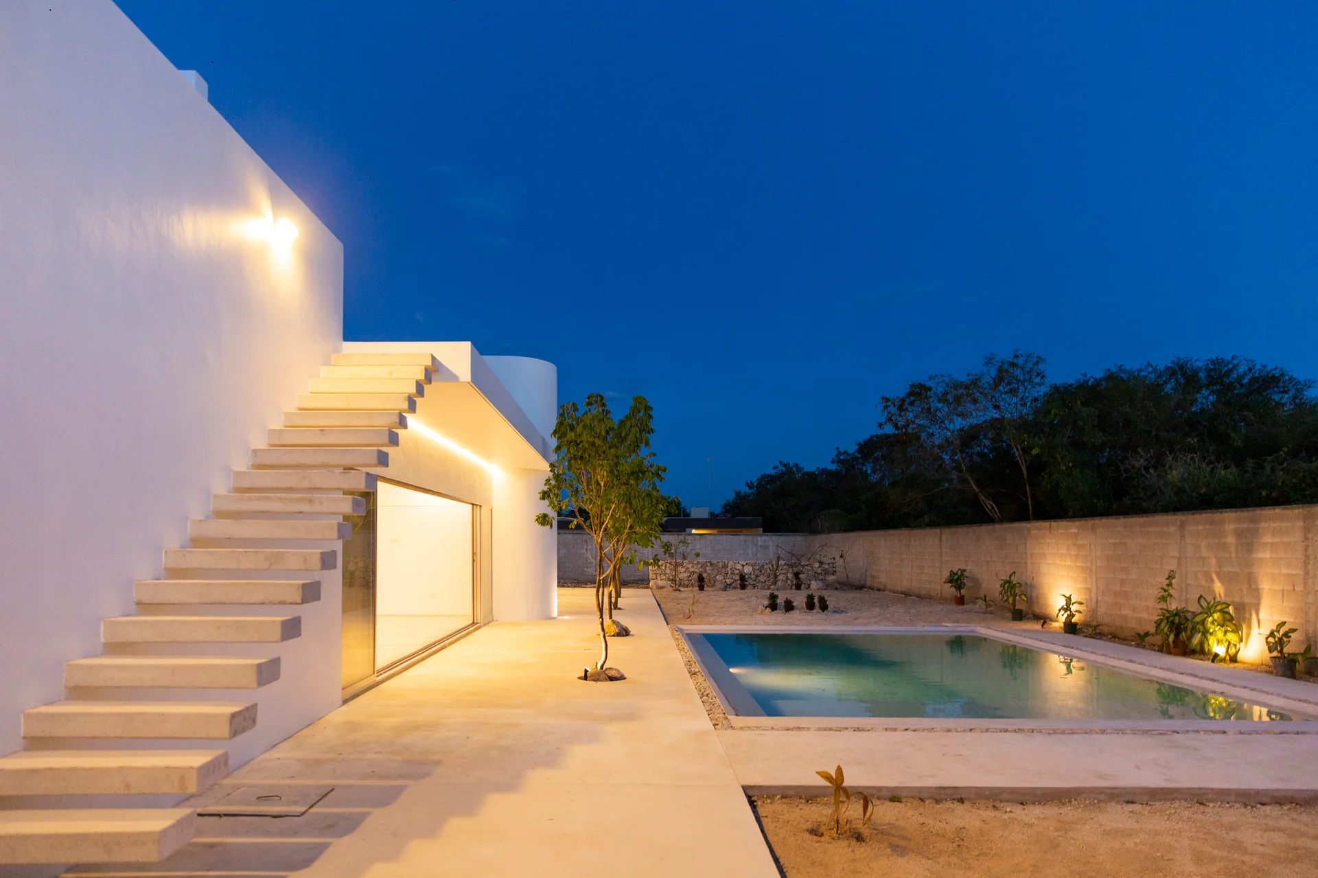 A minimalist white house features a floating concrete staircase, a glowing glass sliding door, and a rectangular swimming pool at dusk.