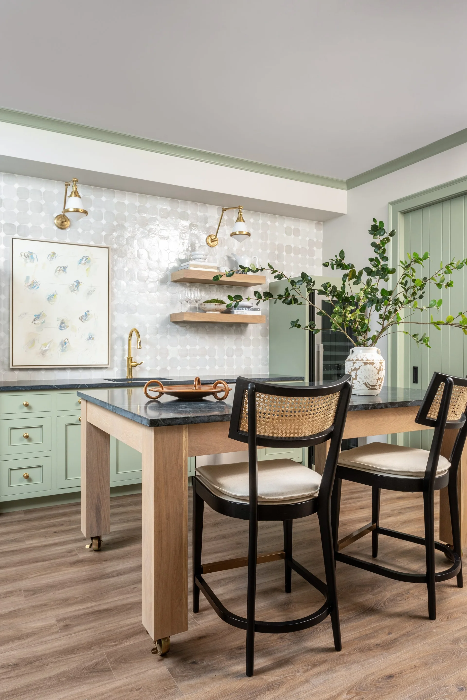 A sage green wet bar area features a hexagon-tiled backsplash, floating wood shelves, and a wooden island with two black cane-back stools.