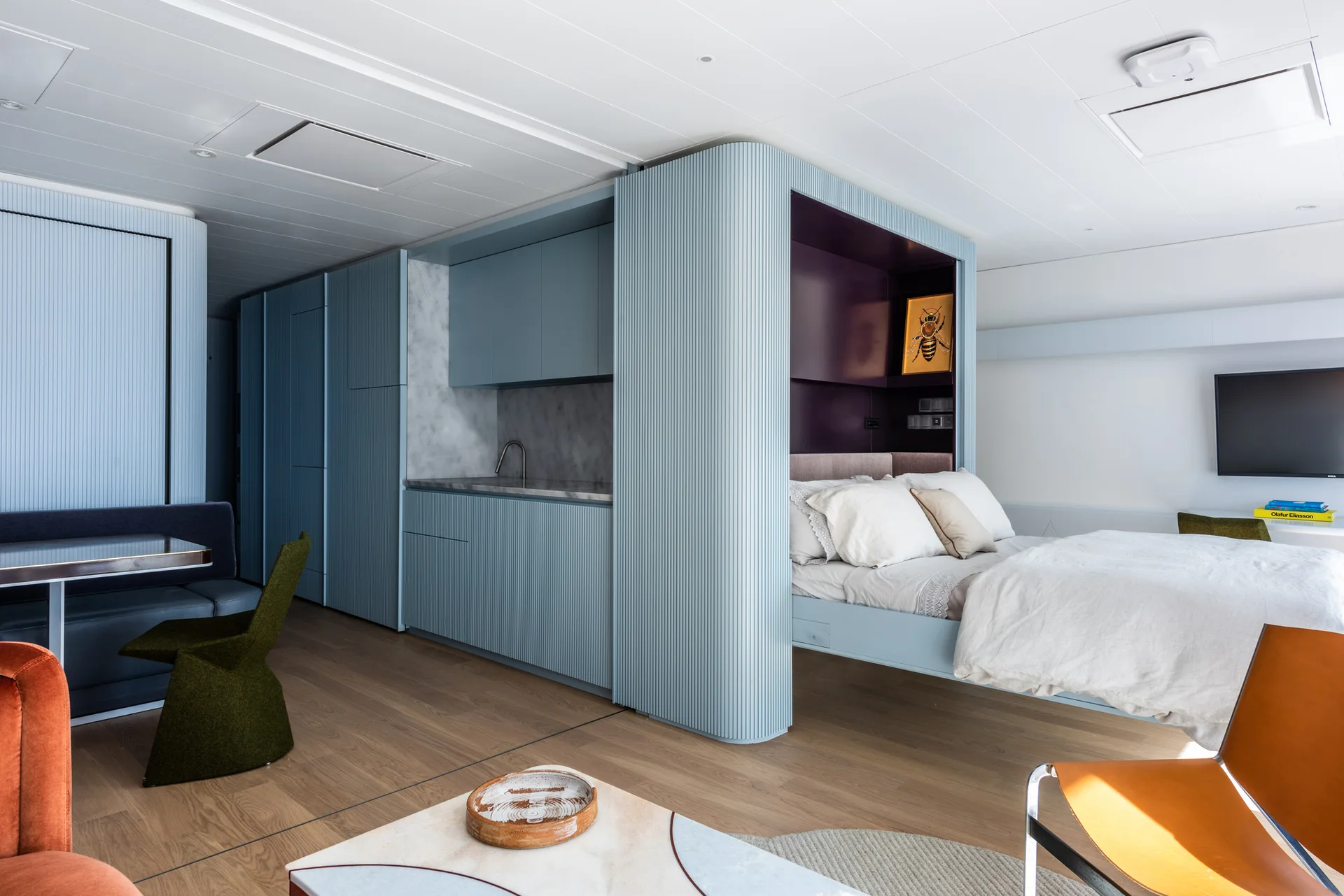 A modern studio apartment interior featuring light blue fluted cabinetry, an integrated kitchenette, and a bed recessed into a deep purple alcove.