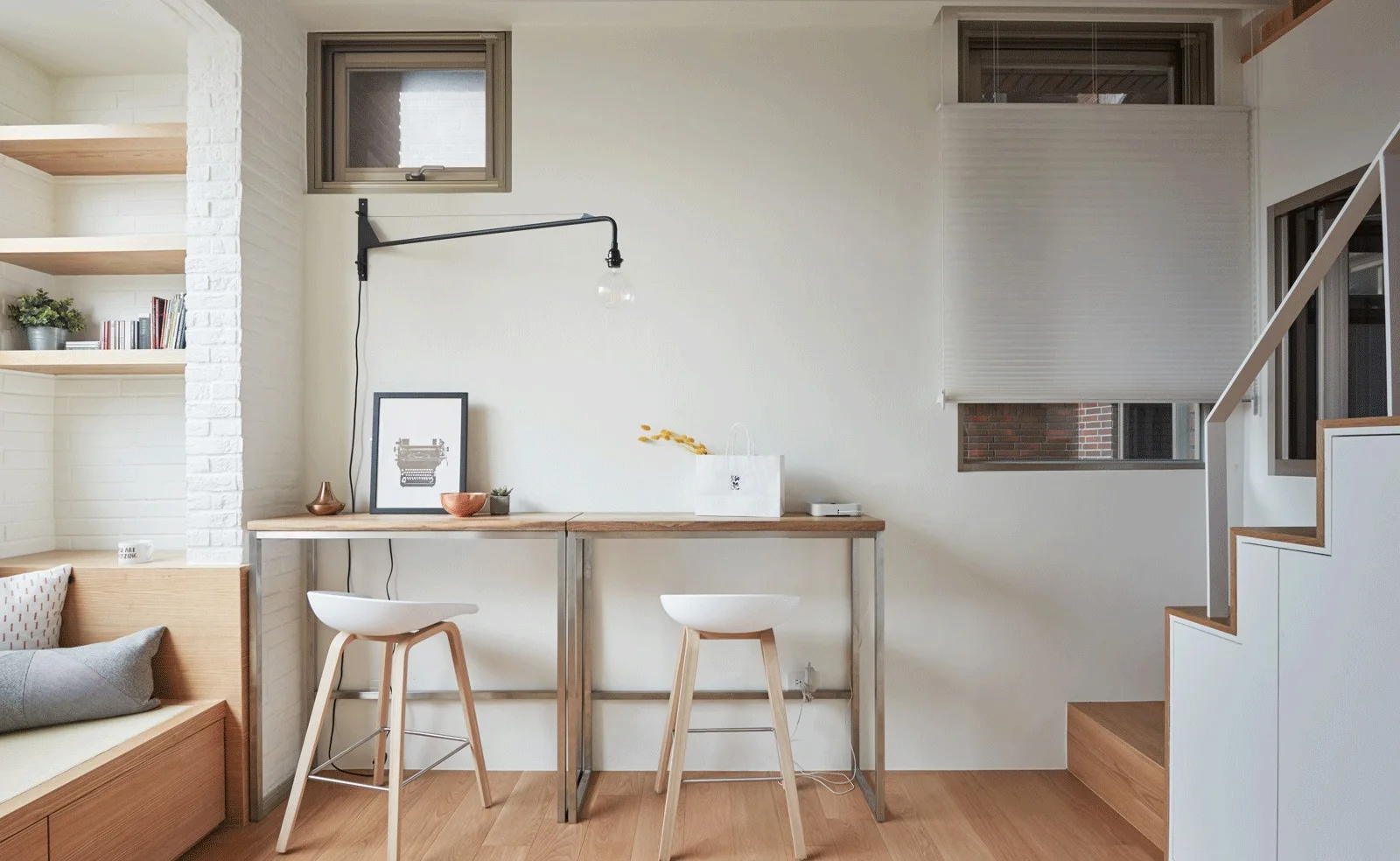 Two wood-topped desks with white counter stools stand in a minimalist workspace featuring floating shelves and a built-in bench.