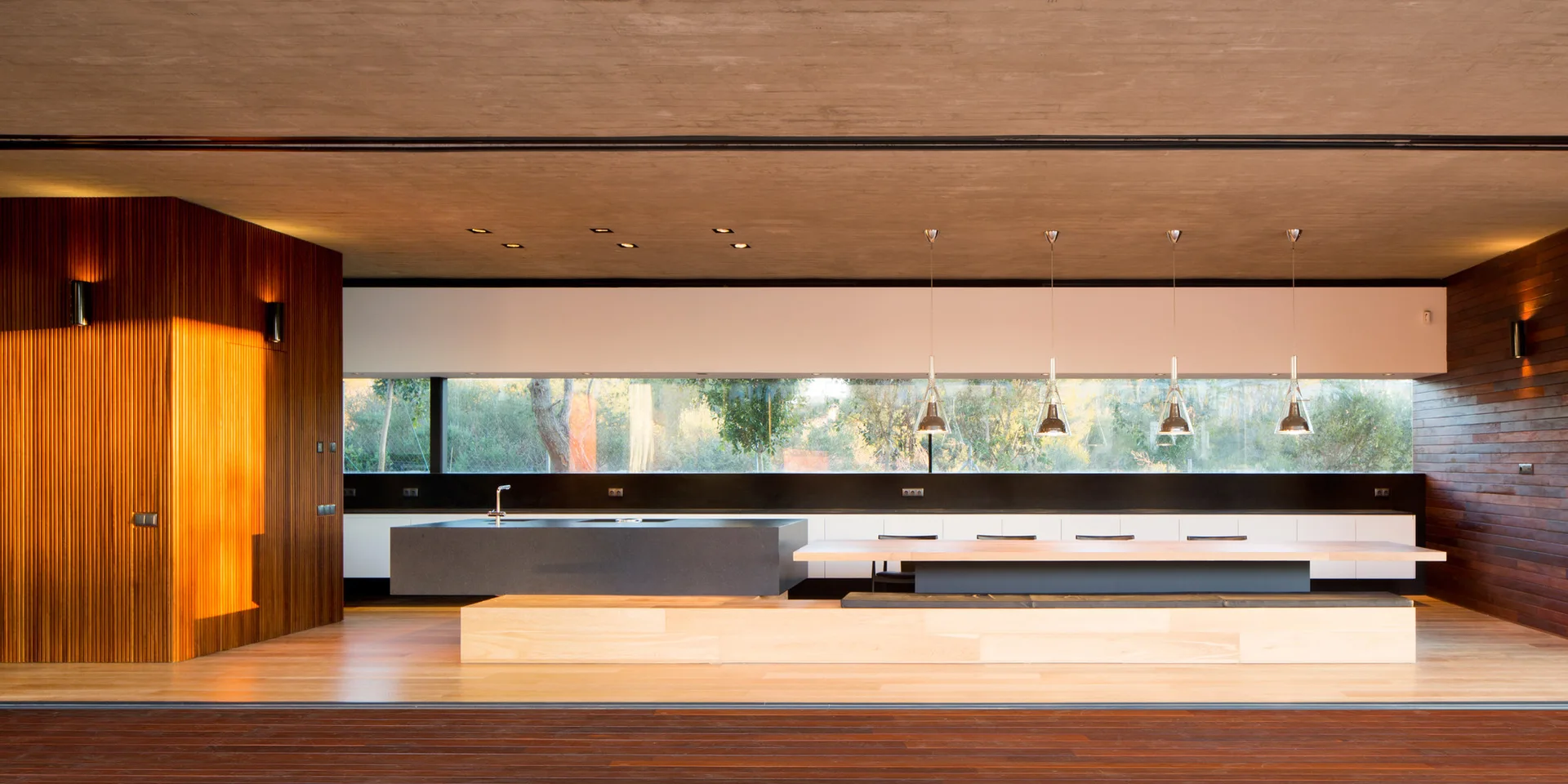 A modern minimalist kitchen featuring a dark grey island with an integrated wood dining table, wood-paneled walls, and a horizontal ribbon window.