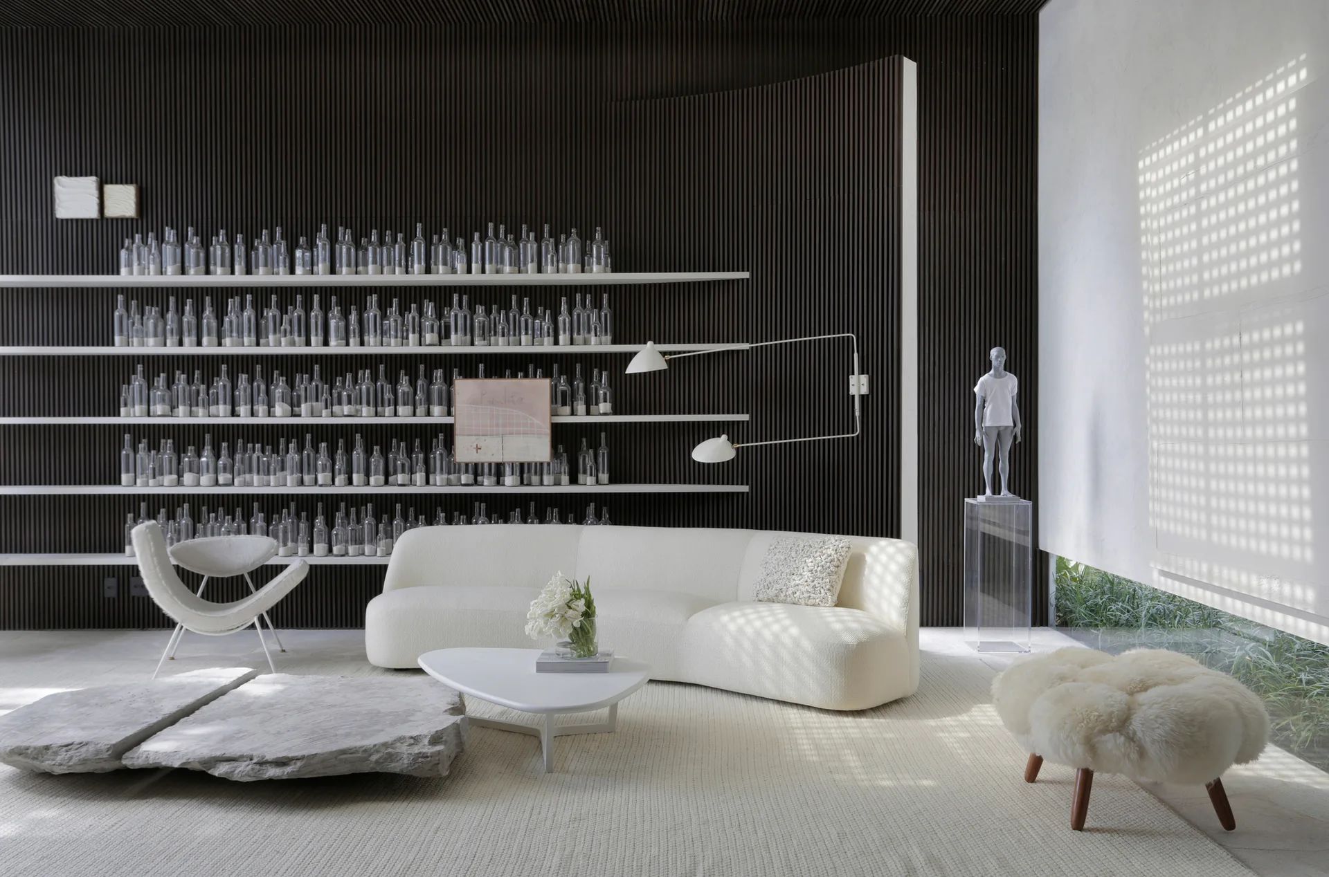 A modern living room featuring a dark slatted accent wall with floating white shelves of glass bottles, a curved white sofa, and a stone coffee table.
