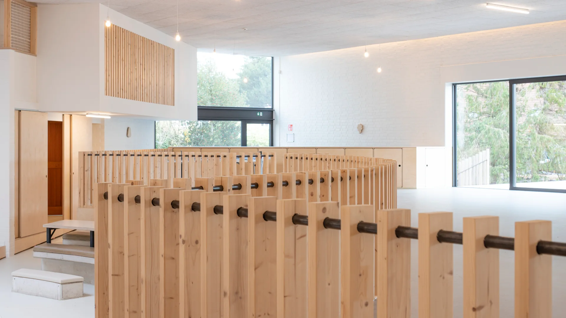 A bright, open space featuring a curved wood slat railing, a white painted brick wall, low wooden cabinetry, and large windows.