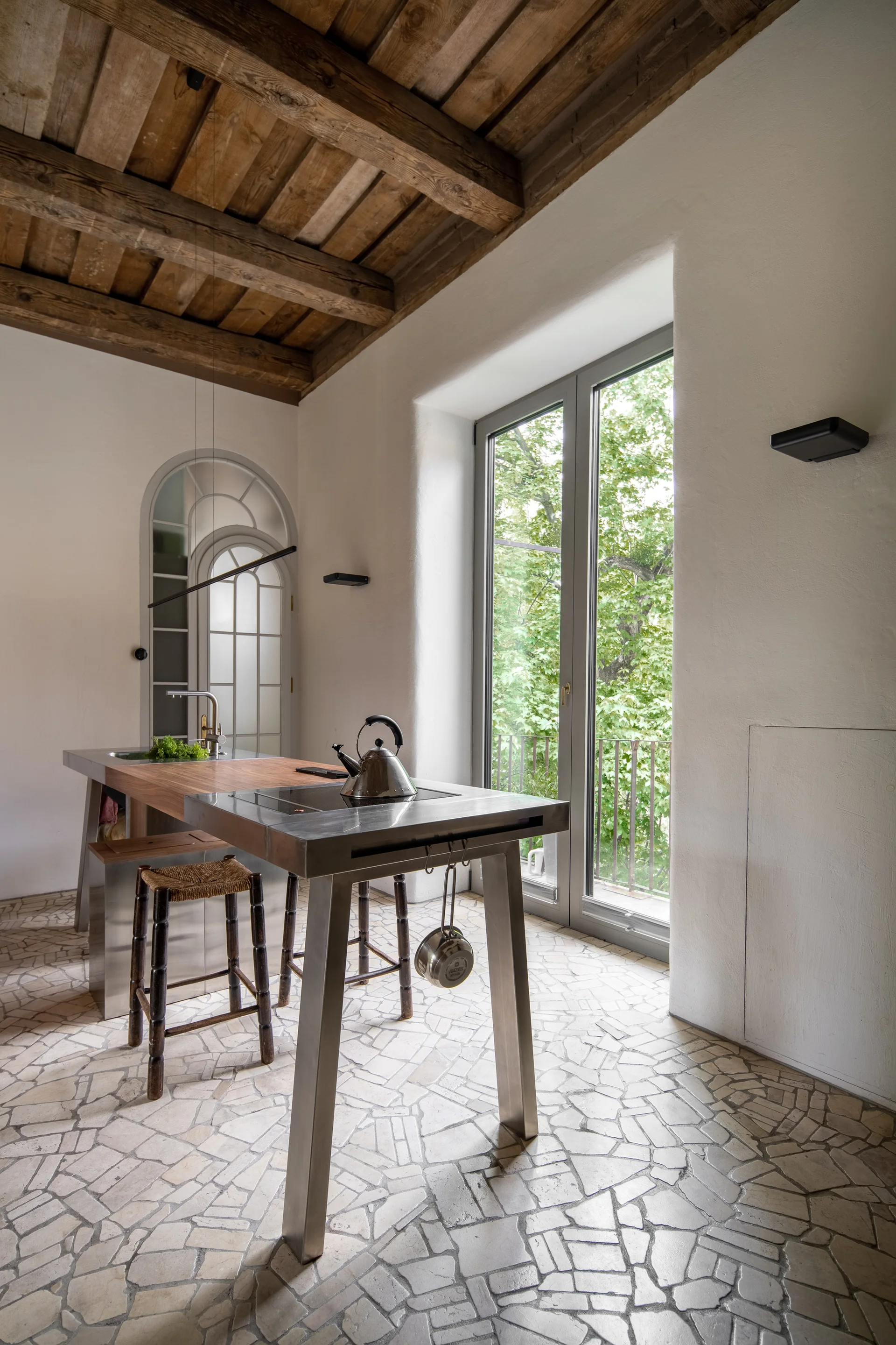 A rustic kitchen features a stainless steel island with a built-in cooktop, exposed wooden ceiling beams, woven stools, and an irregular stone tile floor.