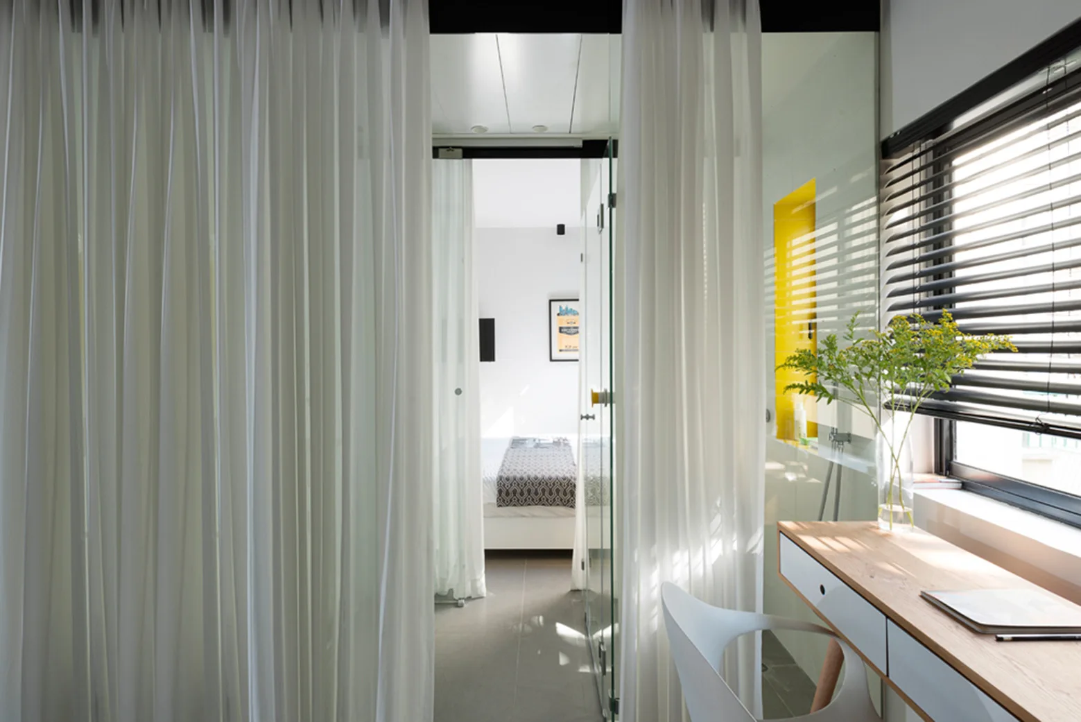 A modern desk and white chair sit beside a window with black blinds, separated from a background bedroom by long sheer white curtains.